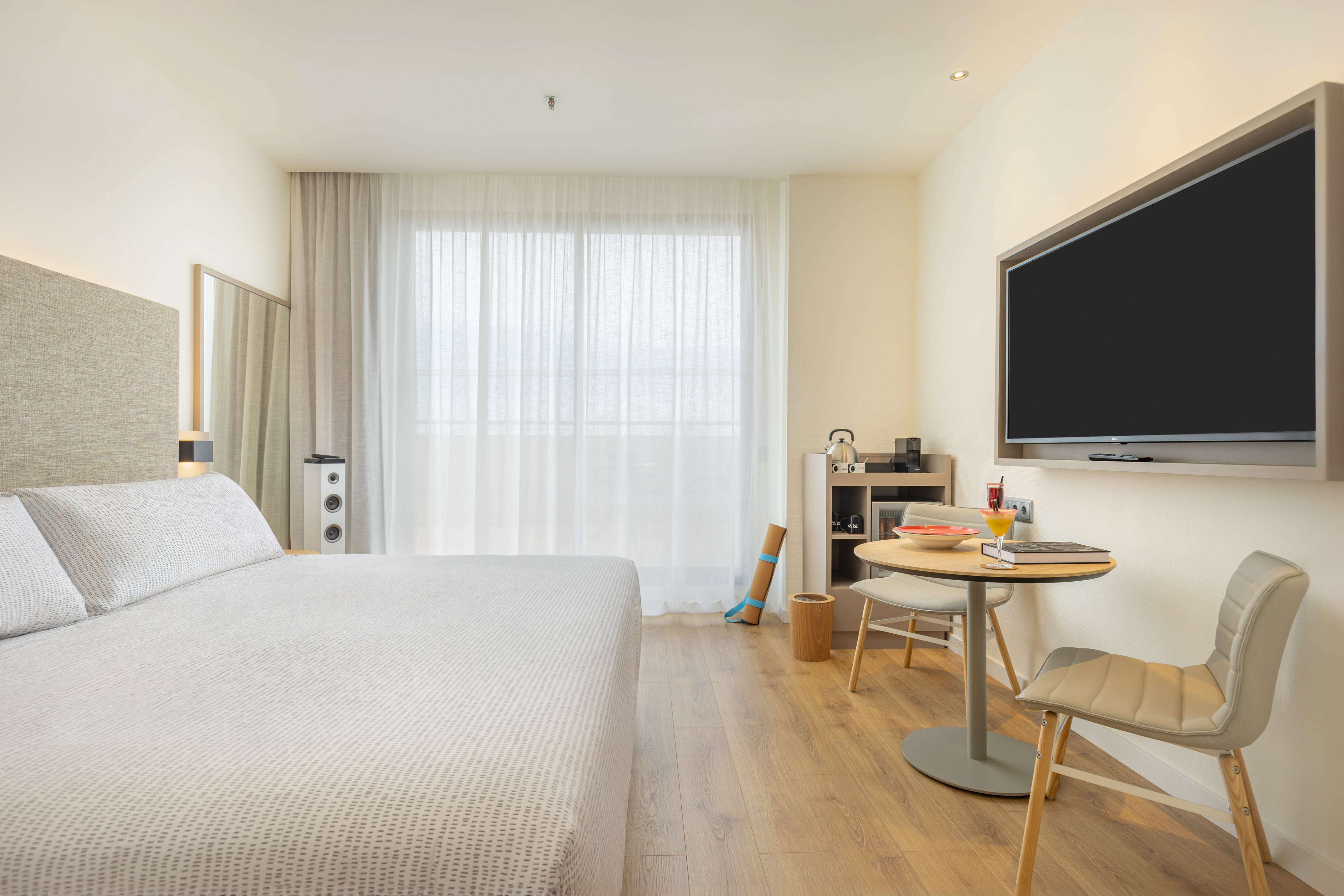 Bright modern hotel room with bed, TV, dining table, and hardwood floor.