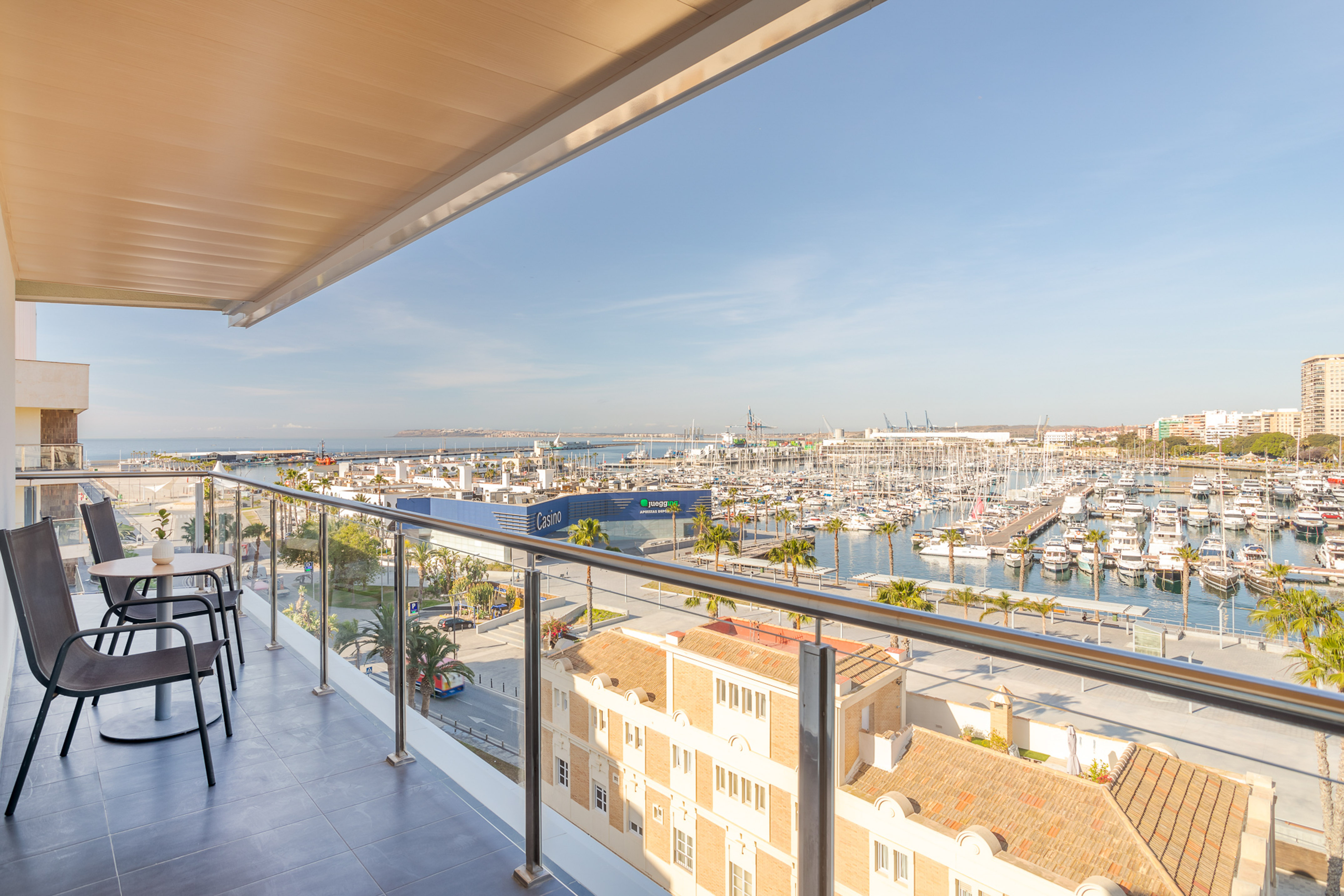Balcony with chairs and awning, overlooking a sunny marina filled with yachts.