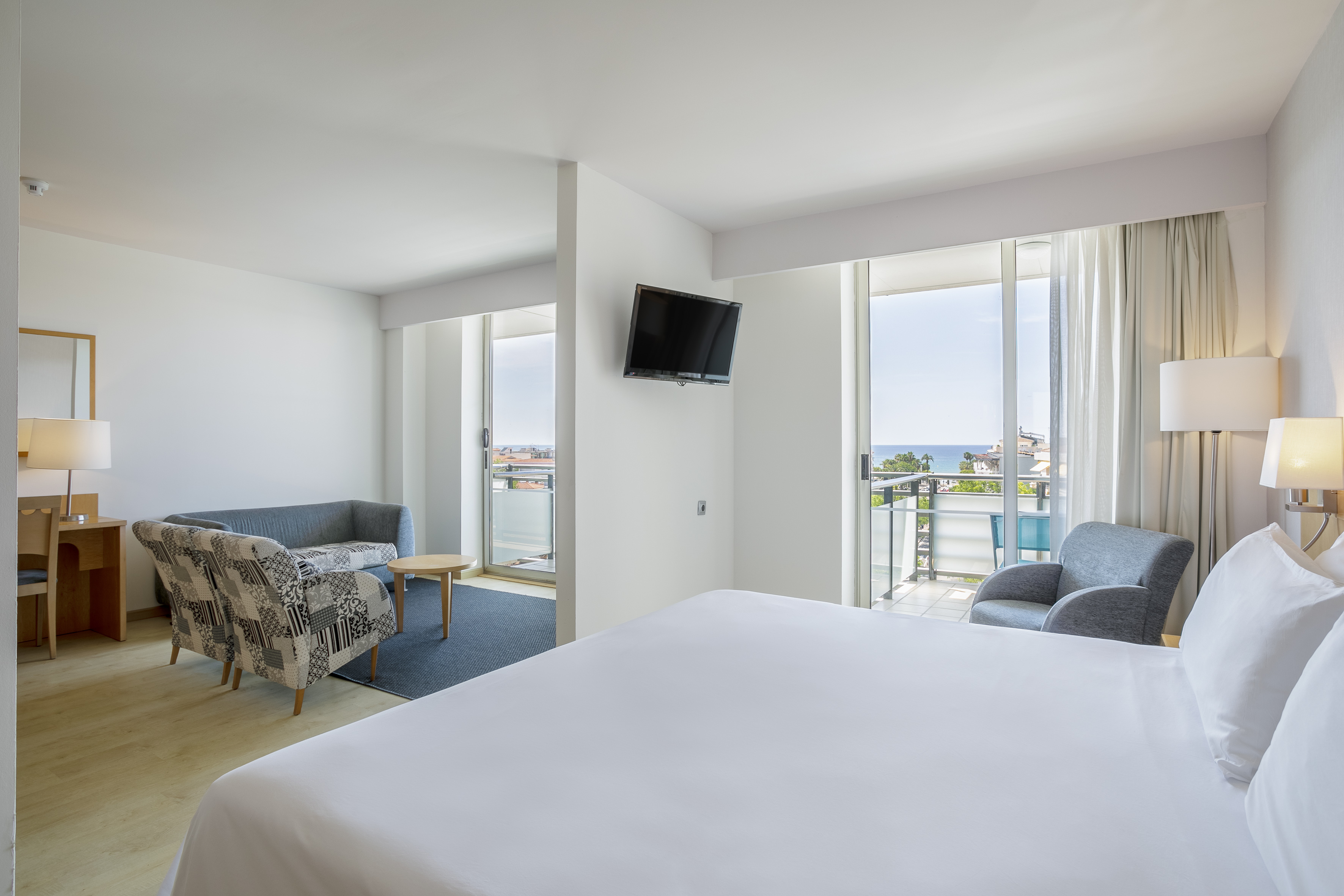 Modern hotel room with ocean view balcony, bed, sofa, and TV.