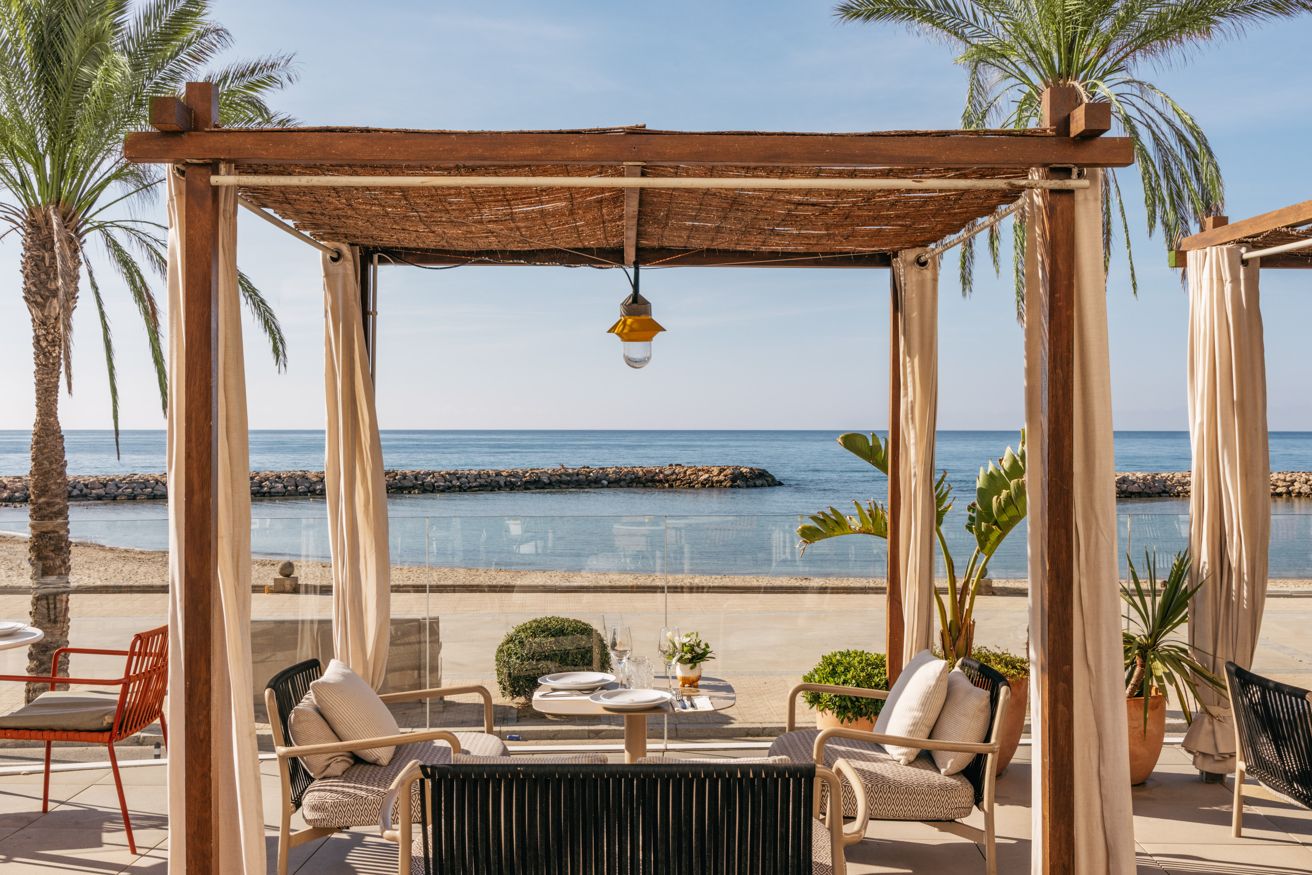 Beachfront dining pergola with table, chairs, palm trees, and ocean view.
