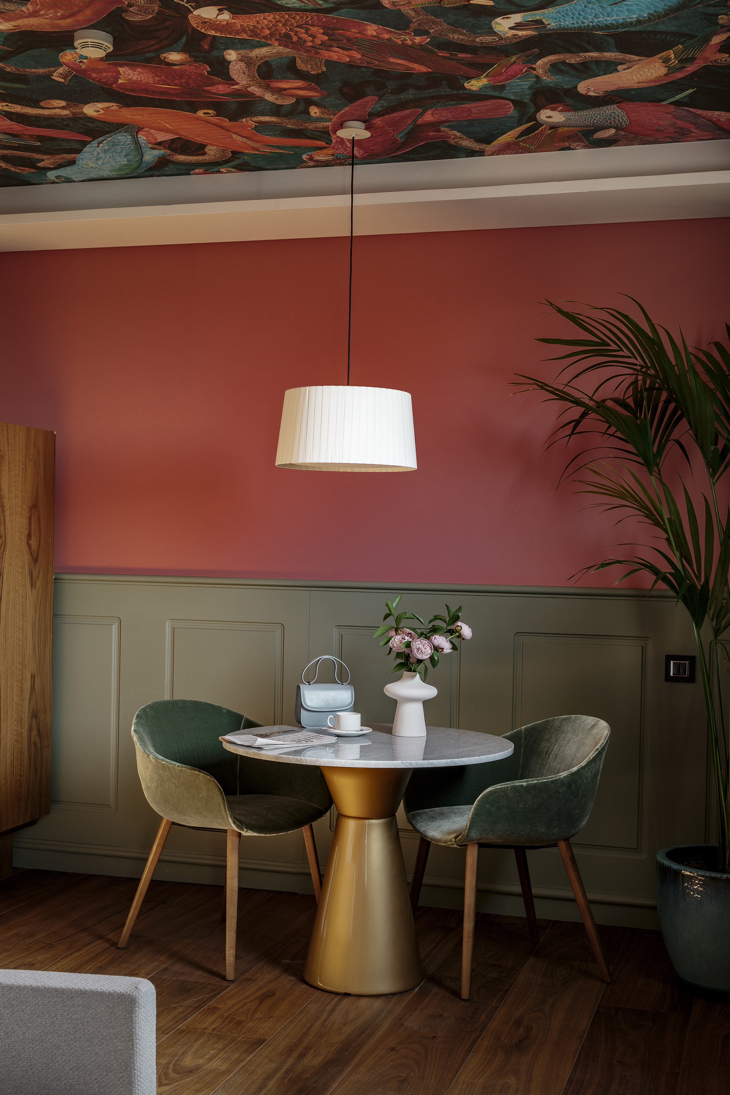 Vibrant room with bird ceiling, coral walls, marble table, green velvet chairs.