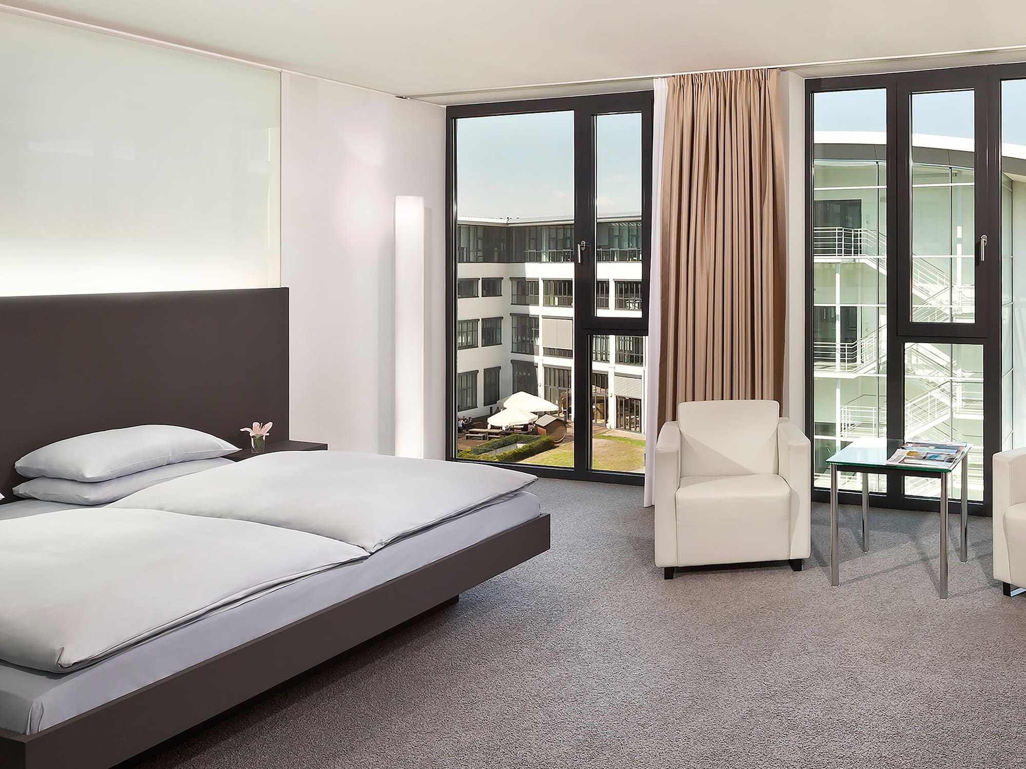 Modern hotel bedroom with large bed, window overlooking courtyard, white armchairs.