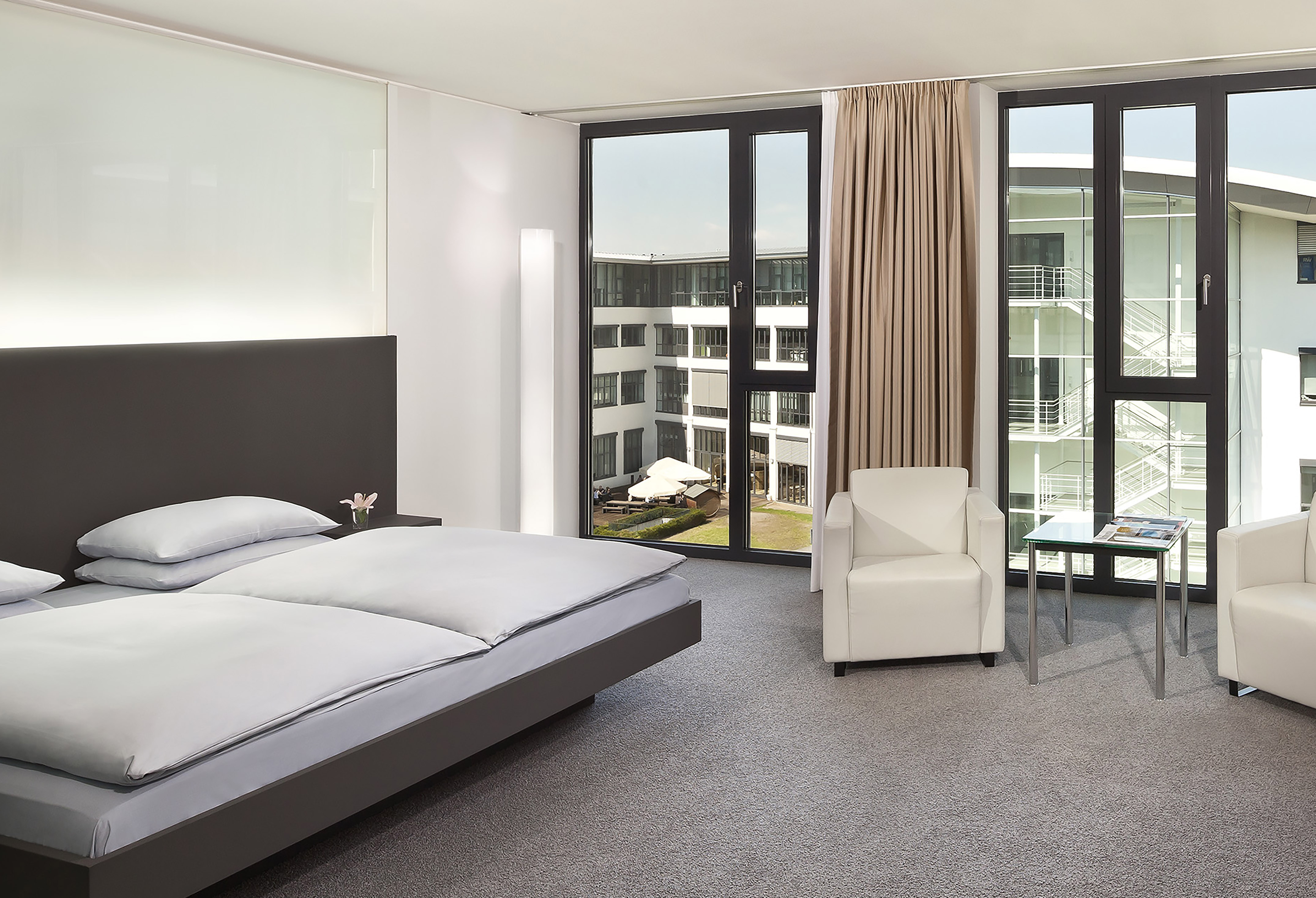 Modern hotel bedroom with large bed, window overlooking courtyard, white armchairs.