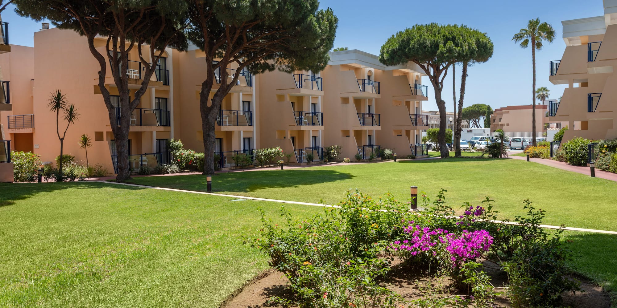 Mediterranean hotel with balconies, green lawn, pine trees, and garden flowers.
