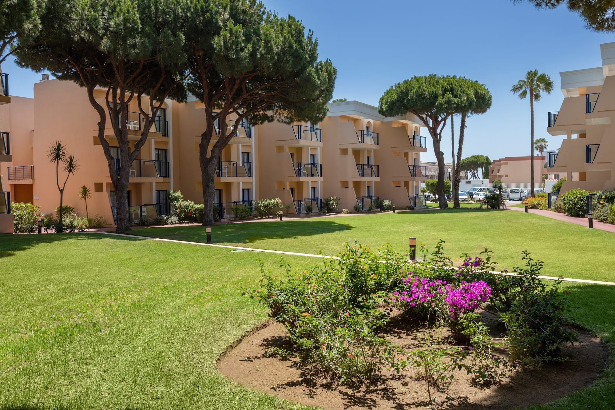 Mediterranean hotel with balconies, green lawn, pine trees, and garden flowers.