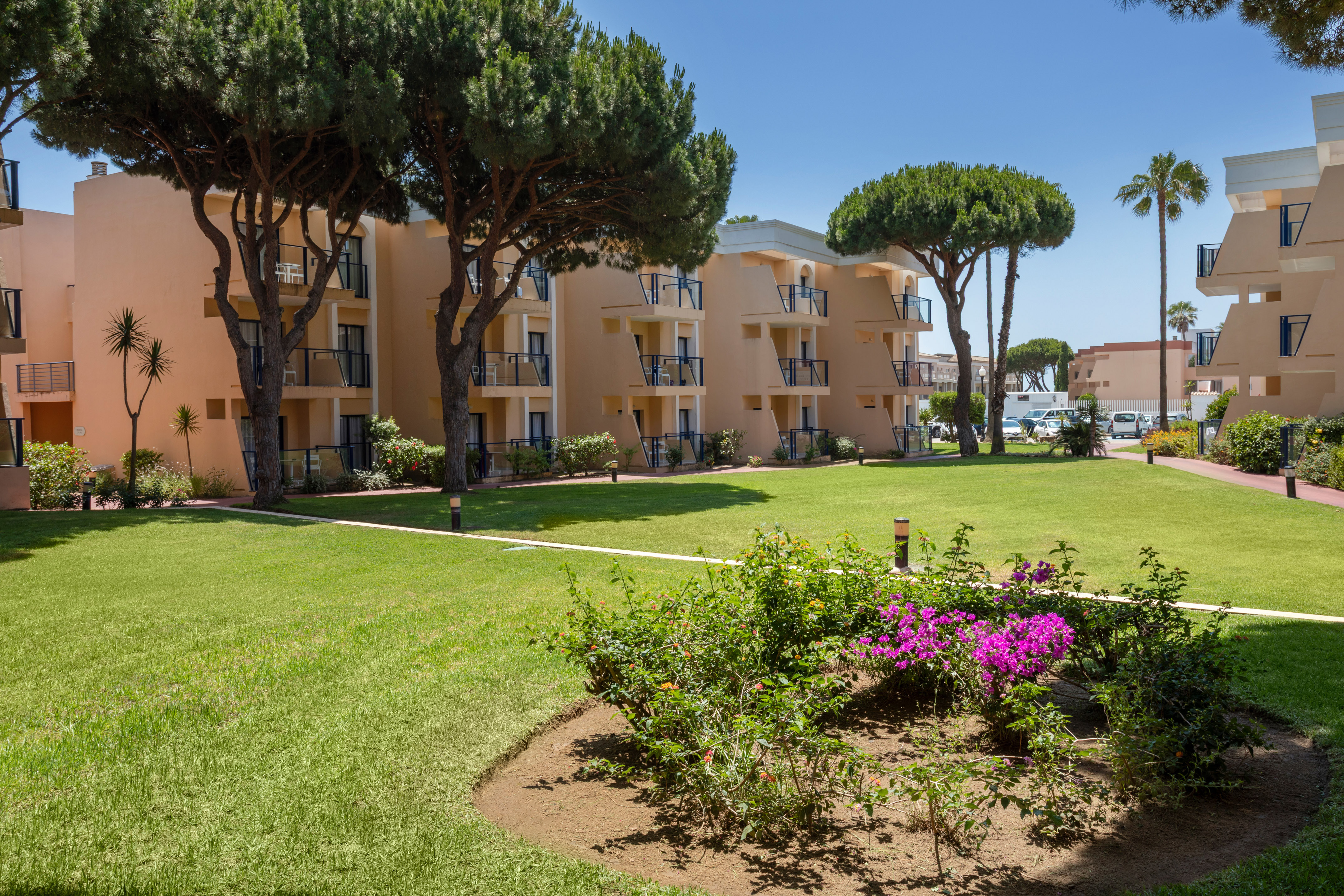 Mediterranean hotel with balconies, green lawn, pine trees, and garden flowers.