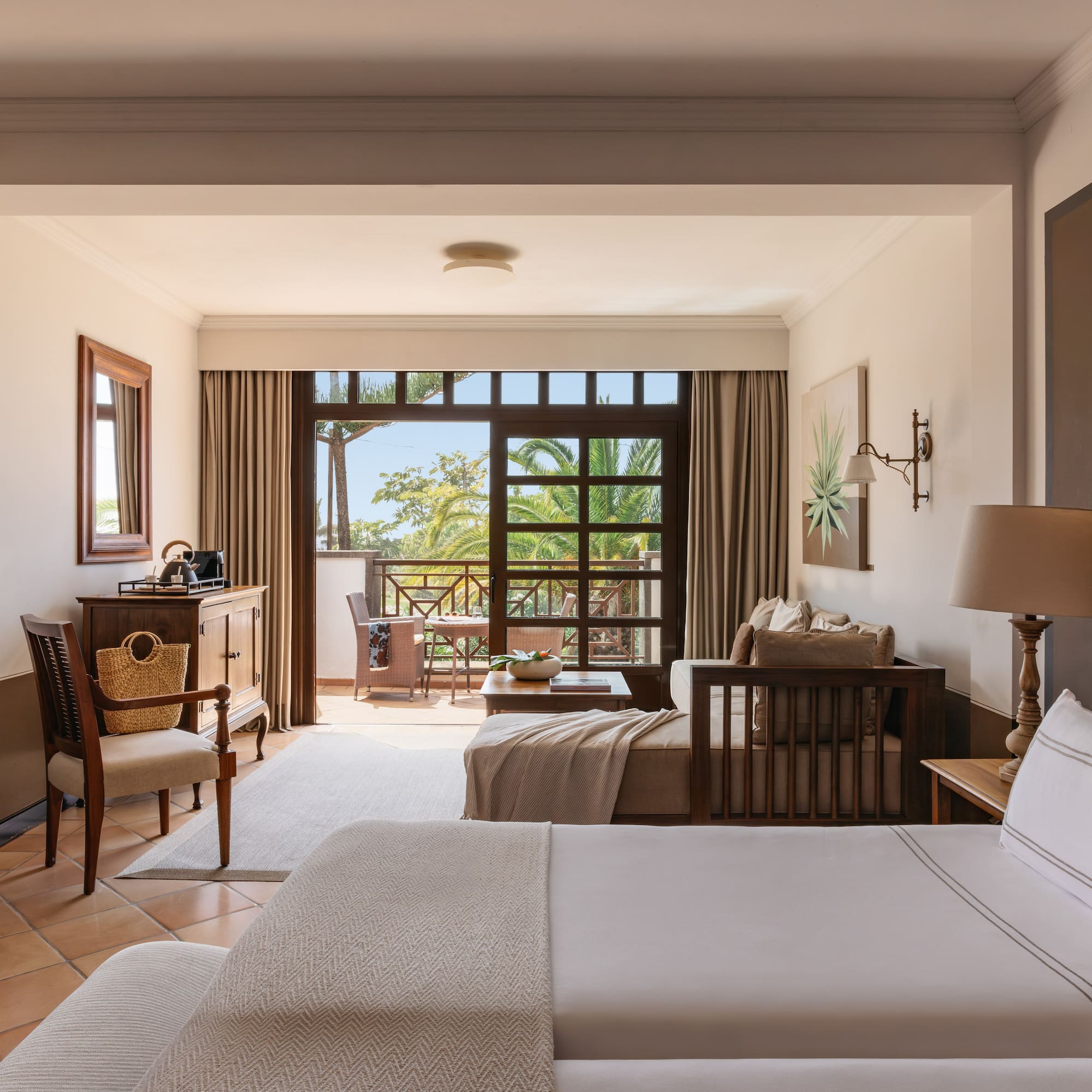 Resort room with bed, sitting area, and balcony overlooking tropical garden.