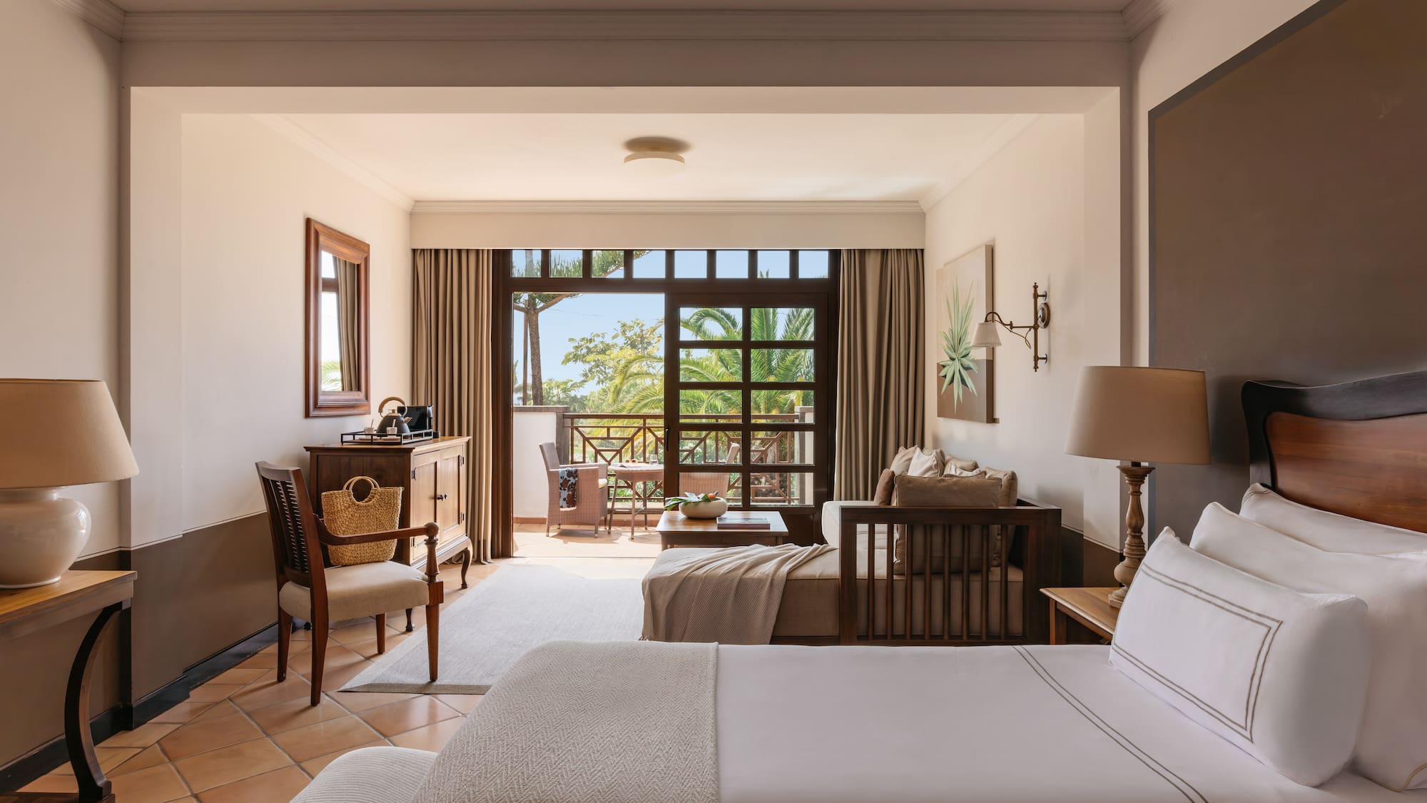 Resort room with bed, sitting area, and balcony overlooking tropical garden.