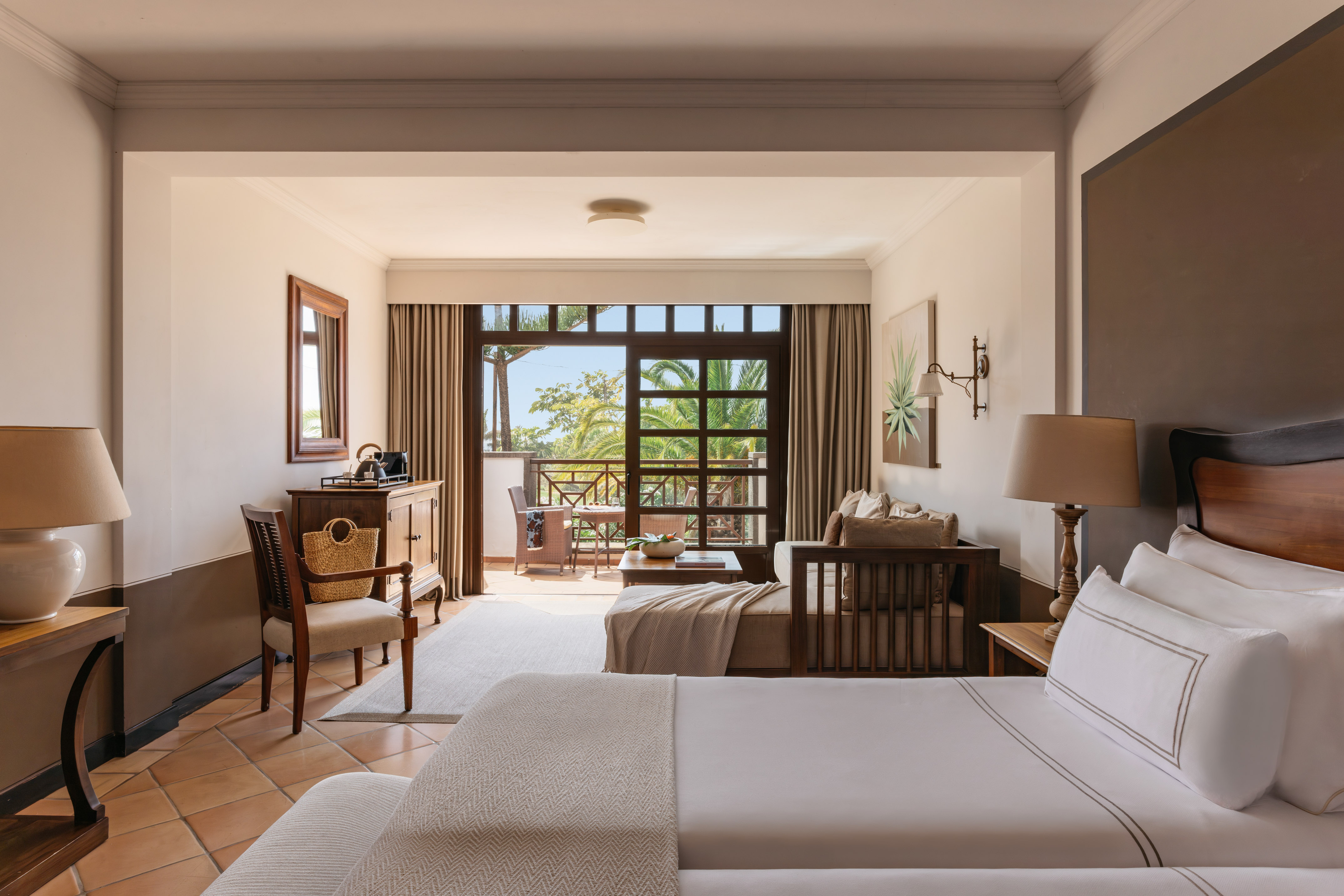Resort room with bed, sitting area, and balcony overlooking tropical garden.