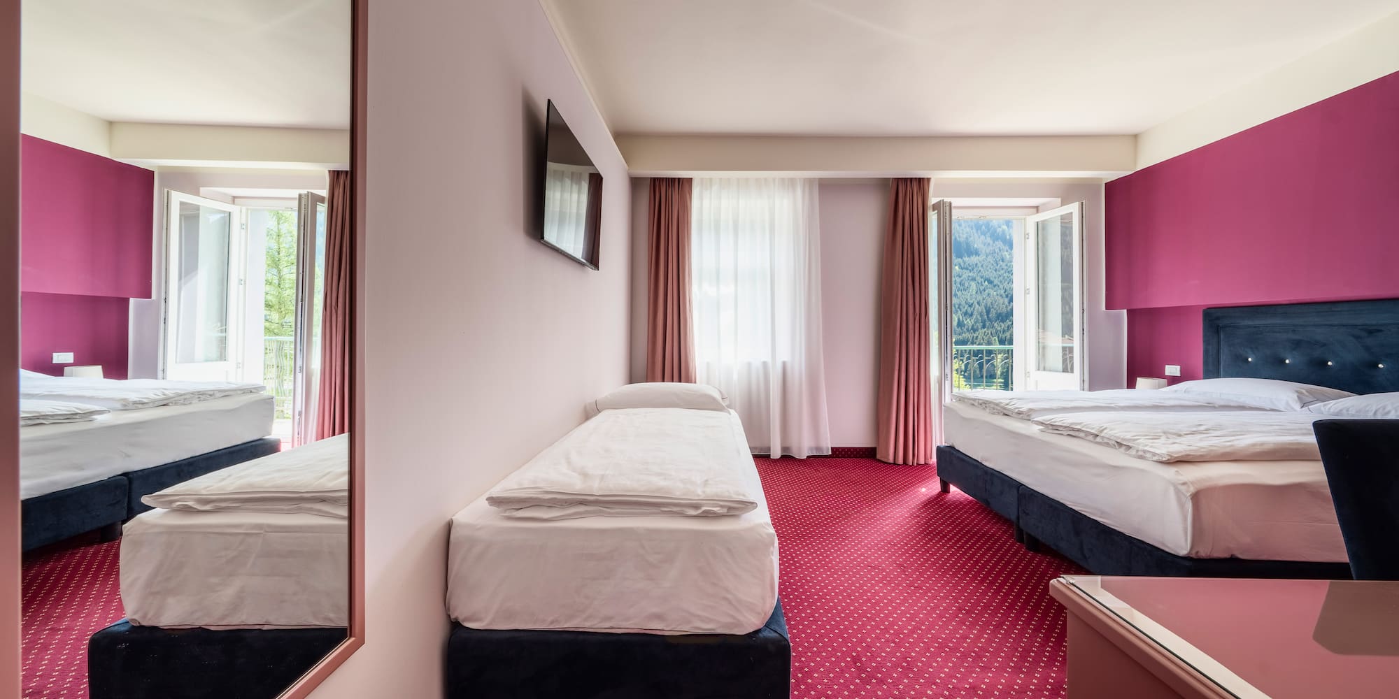 Bright hotel triple room with red carpet, magenta wall, and mountain view.
