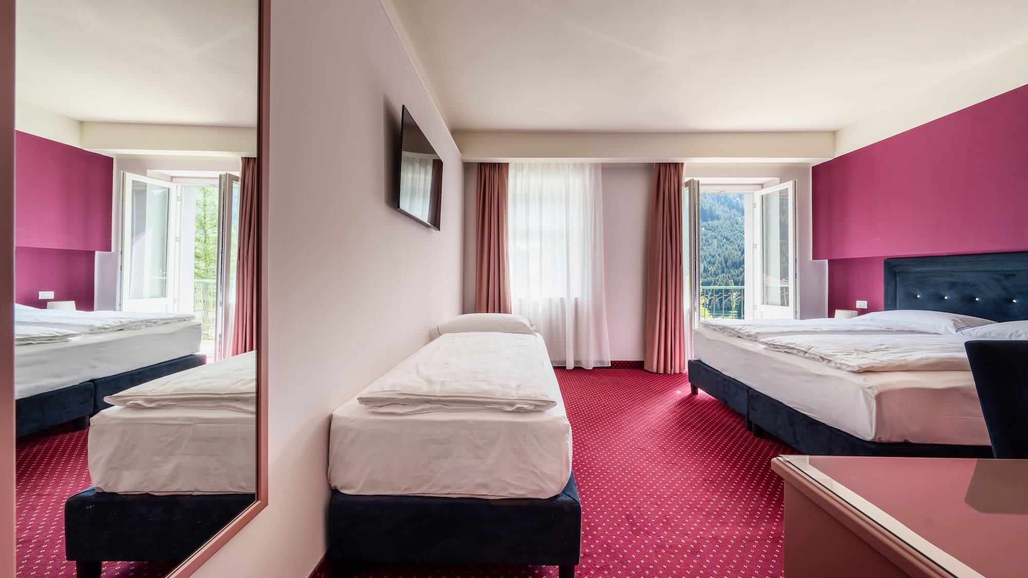 Hotel room with three beds, red carpet, pink walls, mountain view balconies.