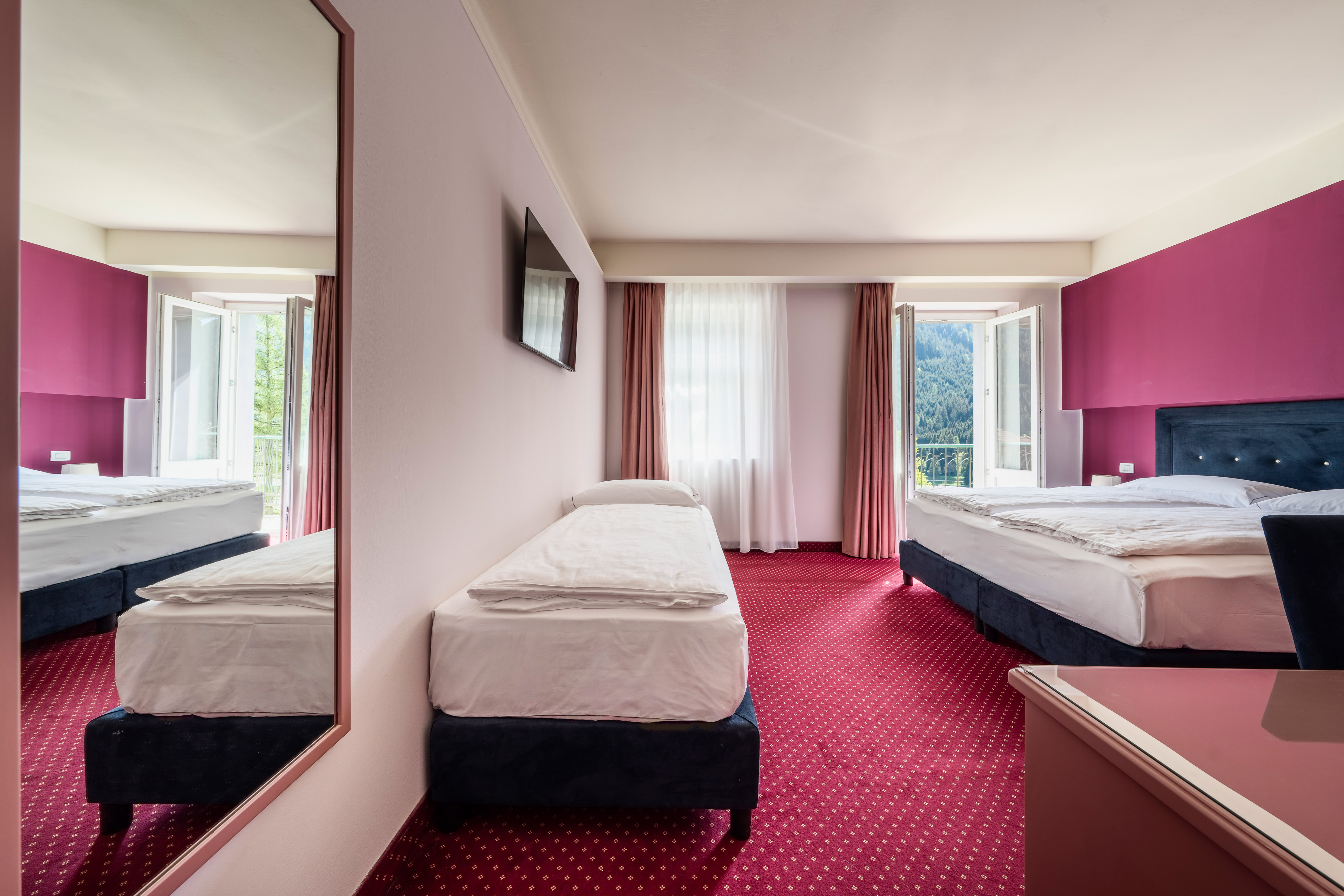 Bright hotel triple room with red carpet, magenta wall, and mountain view.