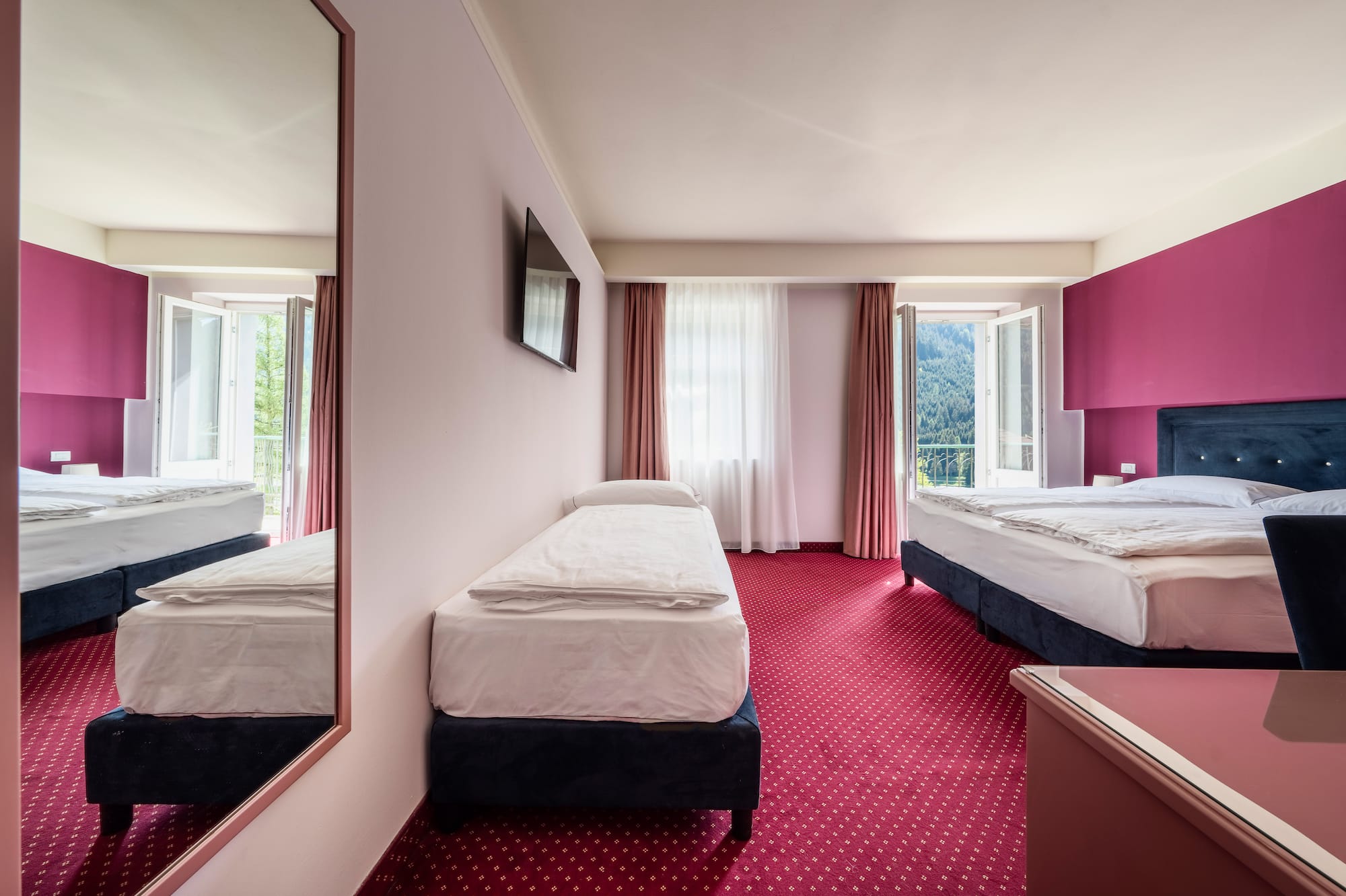 Bright hotel triple room with red carpet, magenta wall, and mountain view.
