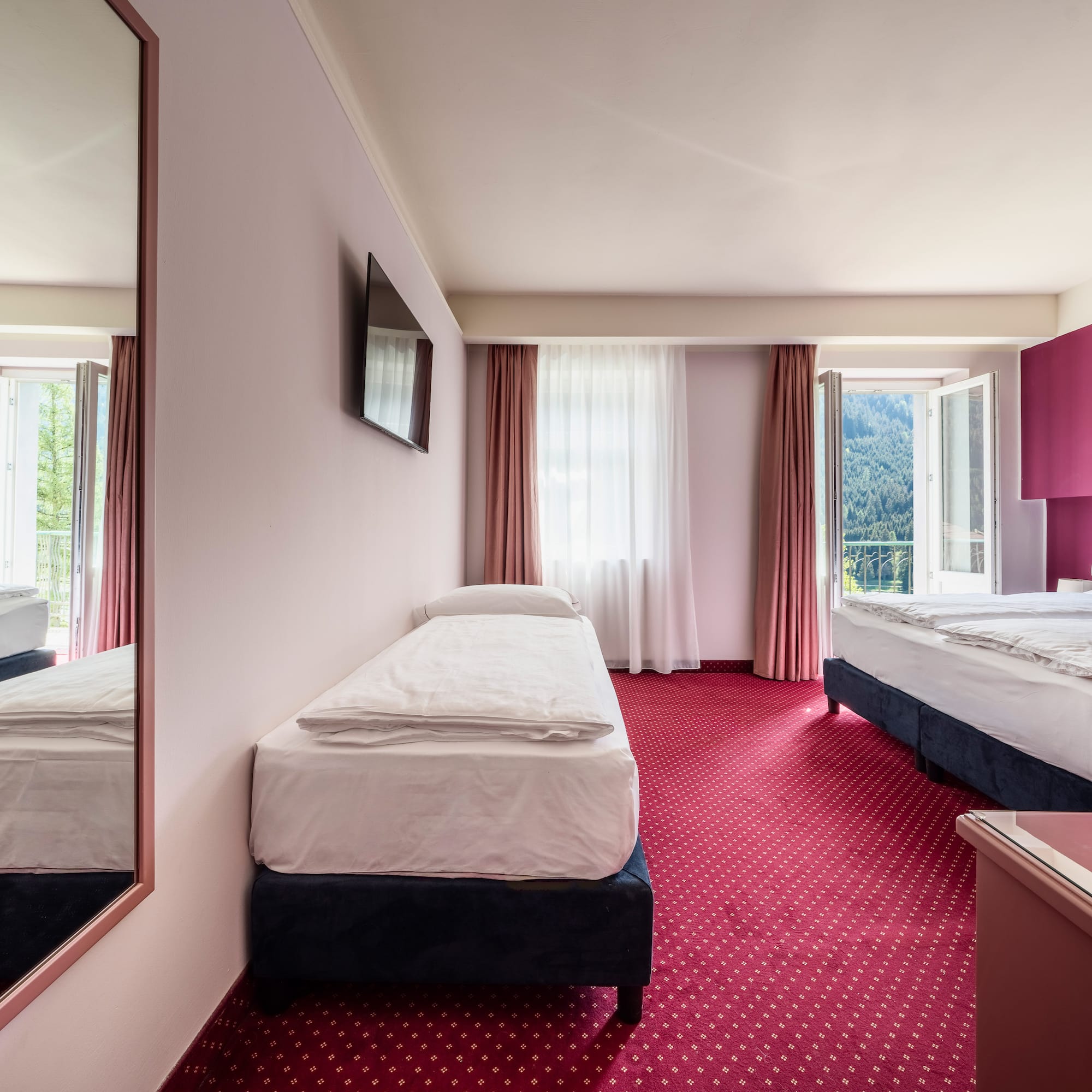 Bright hotel triple room with red carpet, magenta wall, and mountain view.