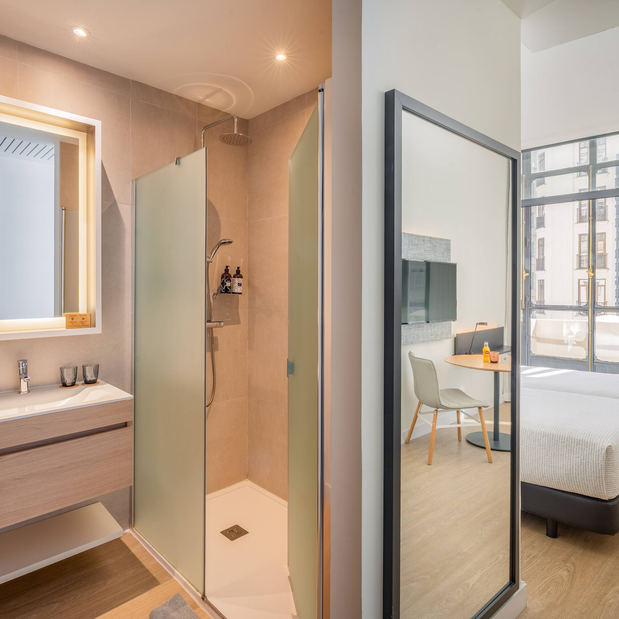 Modern hotel bathroom with walk-in shower, vanity, and twin beds reflected.