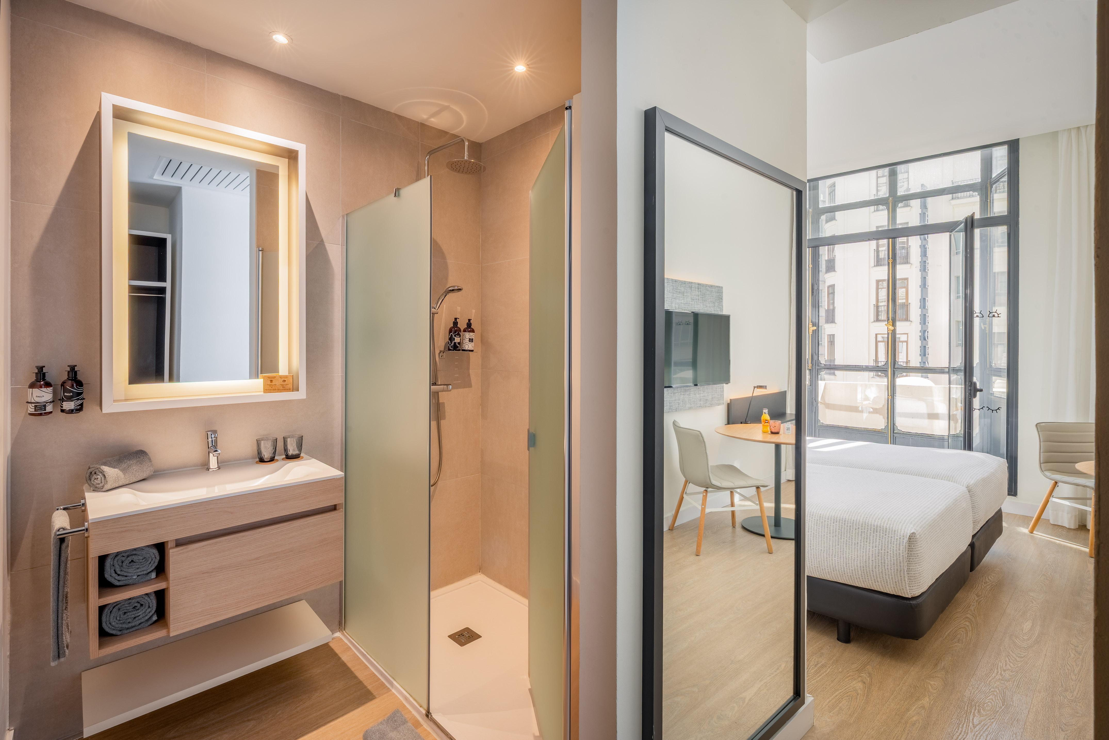Modern hotel bathroom with walk-in shower, vanity, and twin beds reflected.