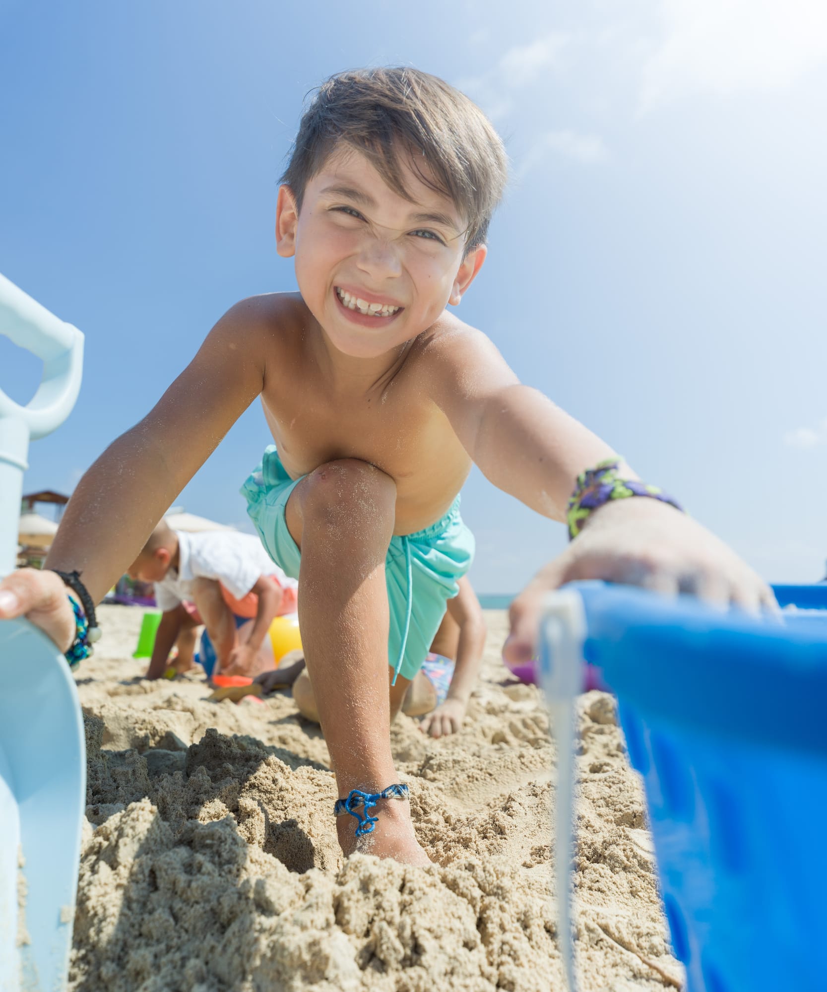 Smiling boy playing on sandy beach with toys under blue sky.