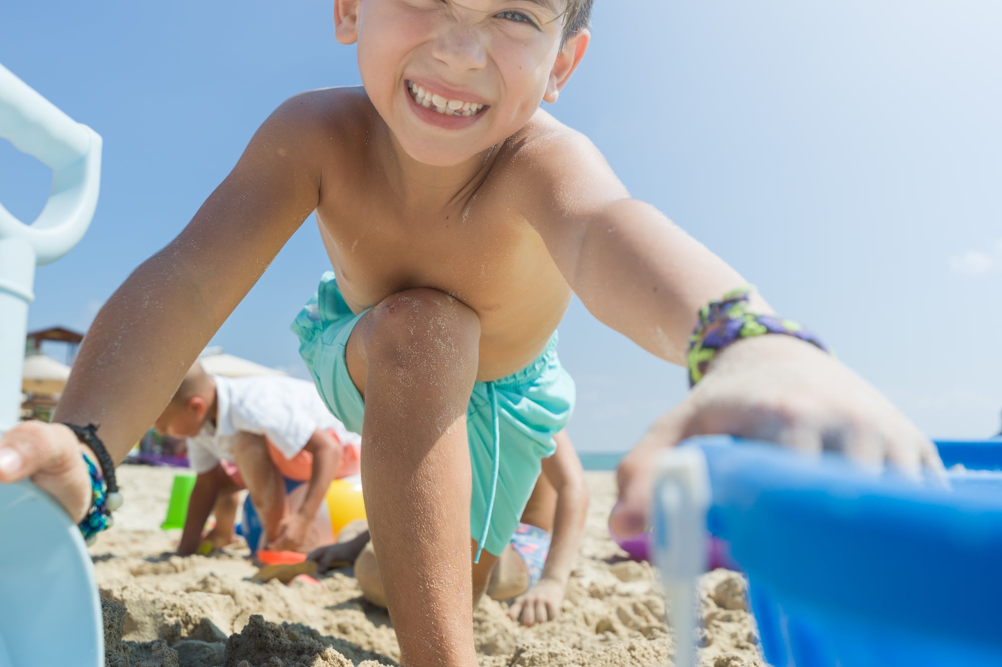 Smiling boy playing on sandy beach with toys under blue sky.