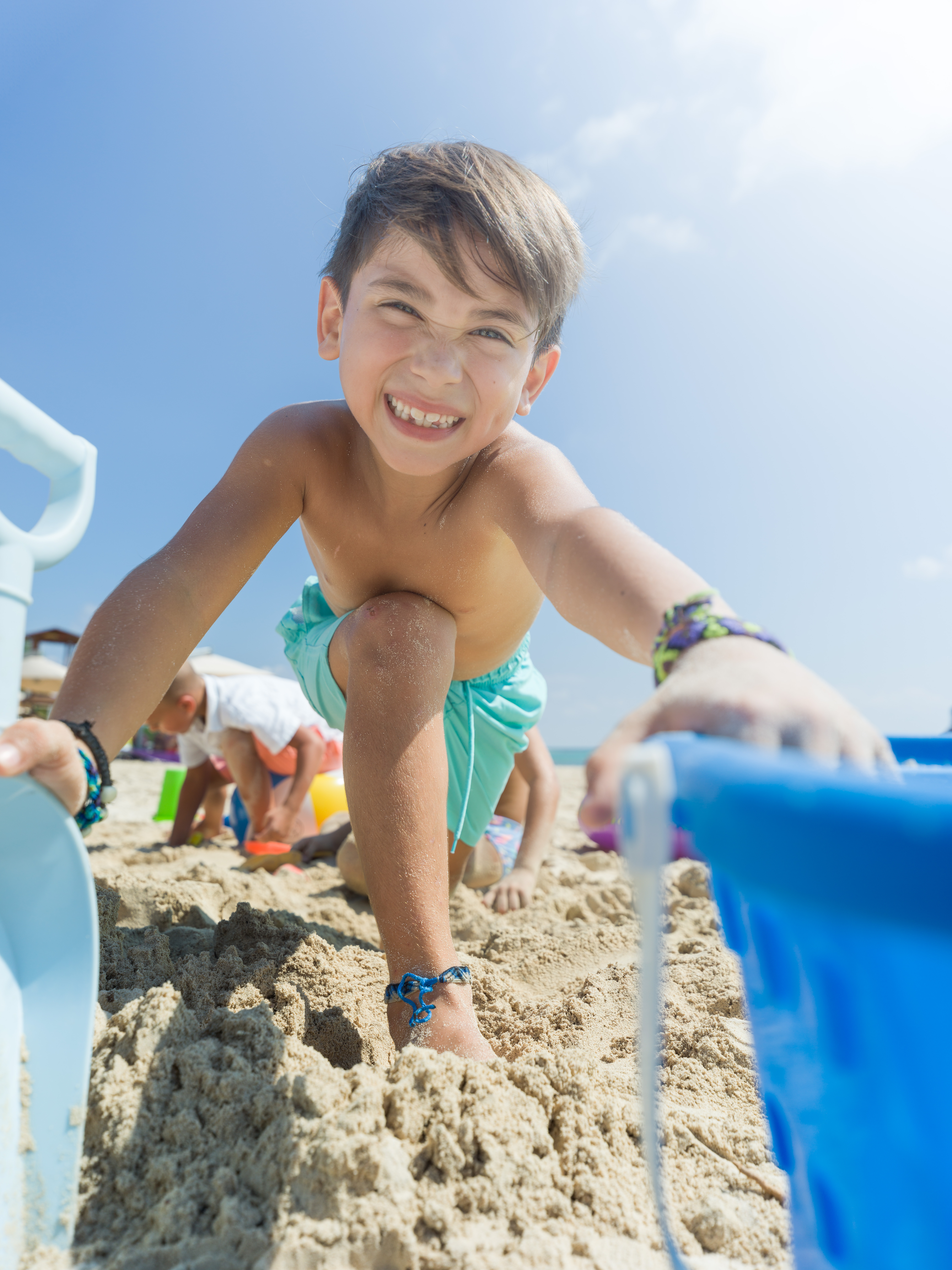 Smiling boy playing on sandy beach with toys under blue sky.