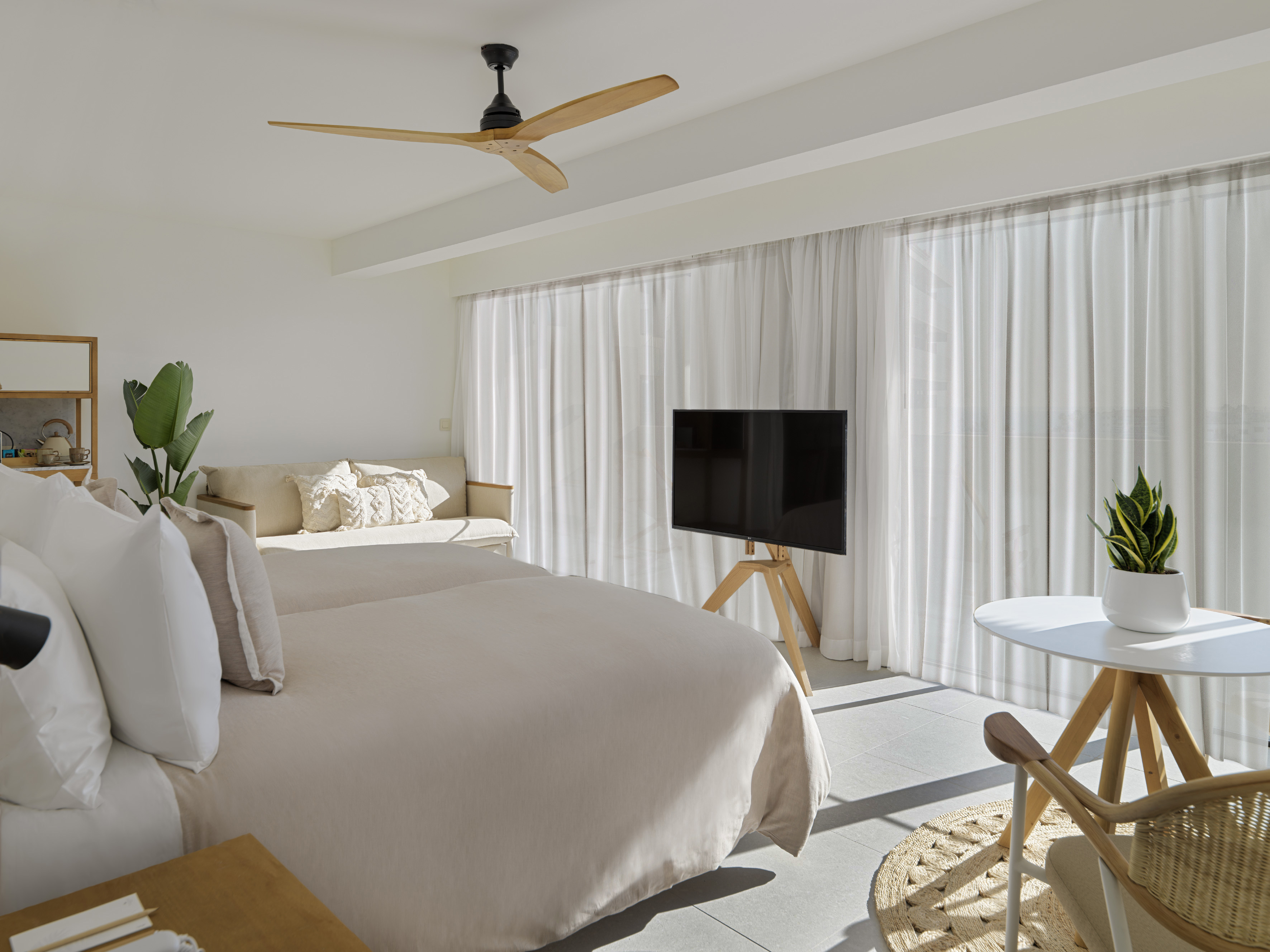 Modern bedroom with two beds, ceiling fan, TV, and bright sheer curtains.