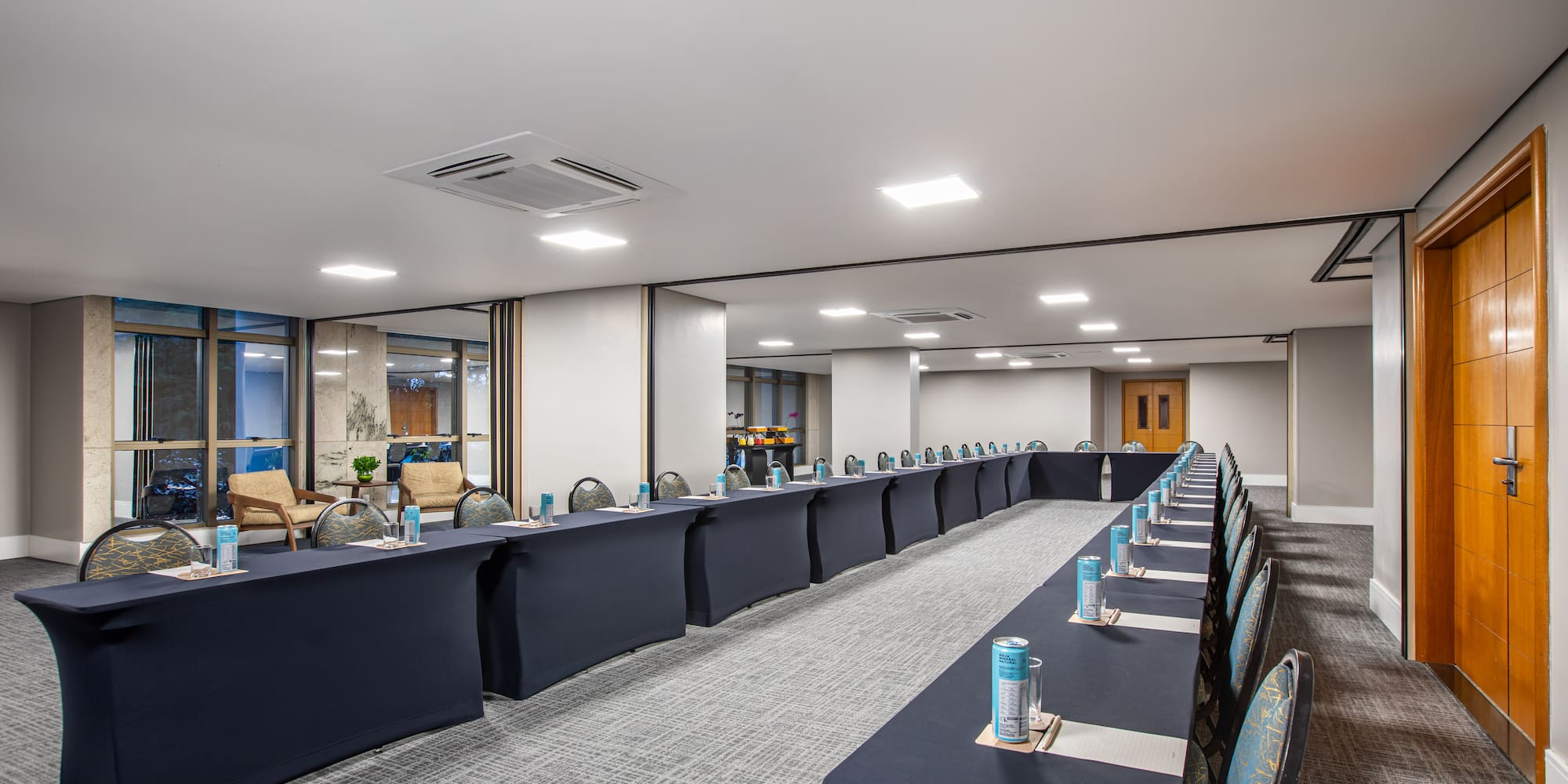 U-shaped meeting room setup with dark tables, chairs, individual drinks, and natural light.