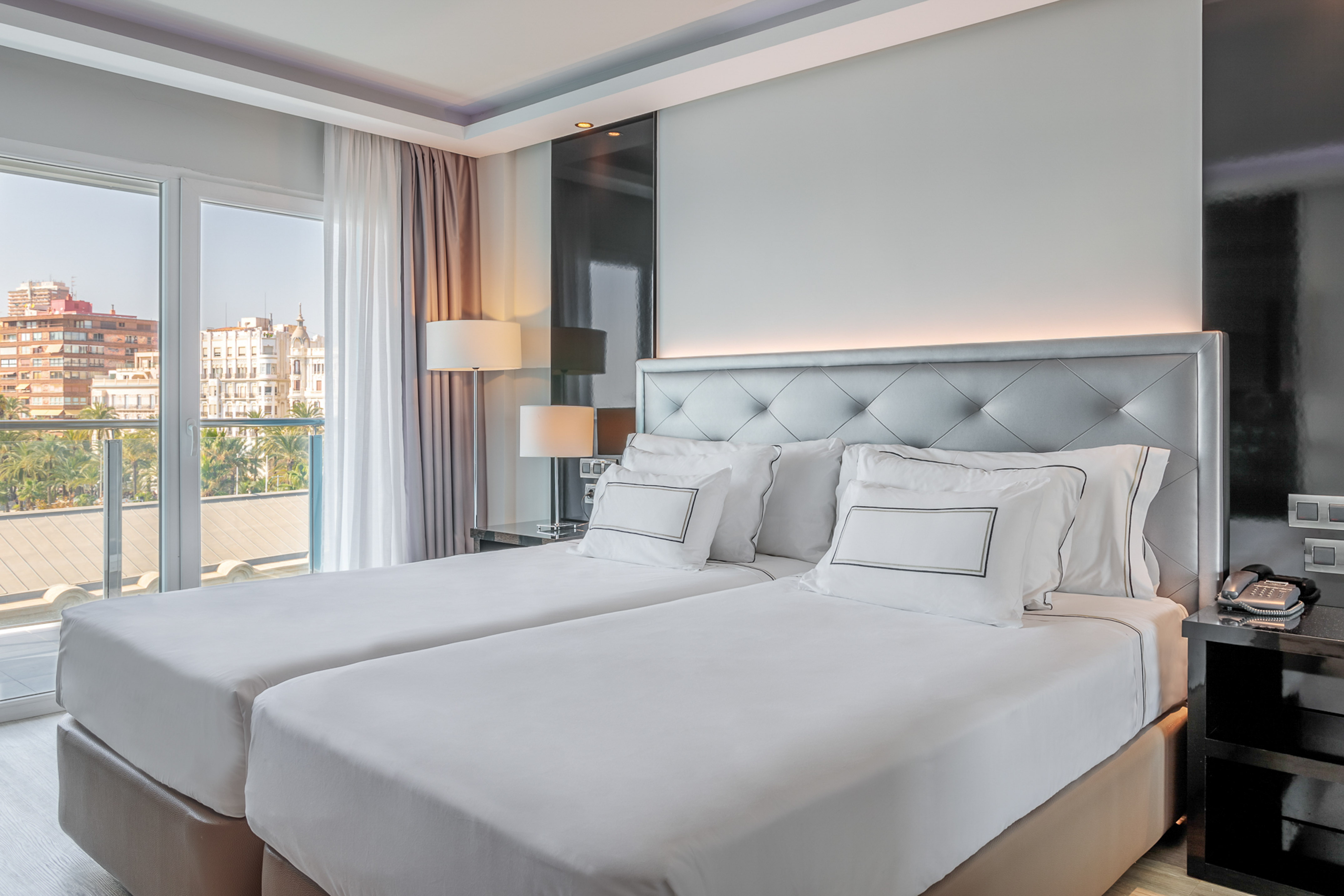 Modern hotel room with twin beds, tufted headboard, and city balcony view.