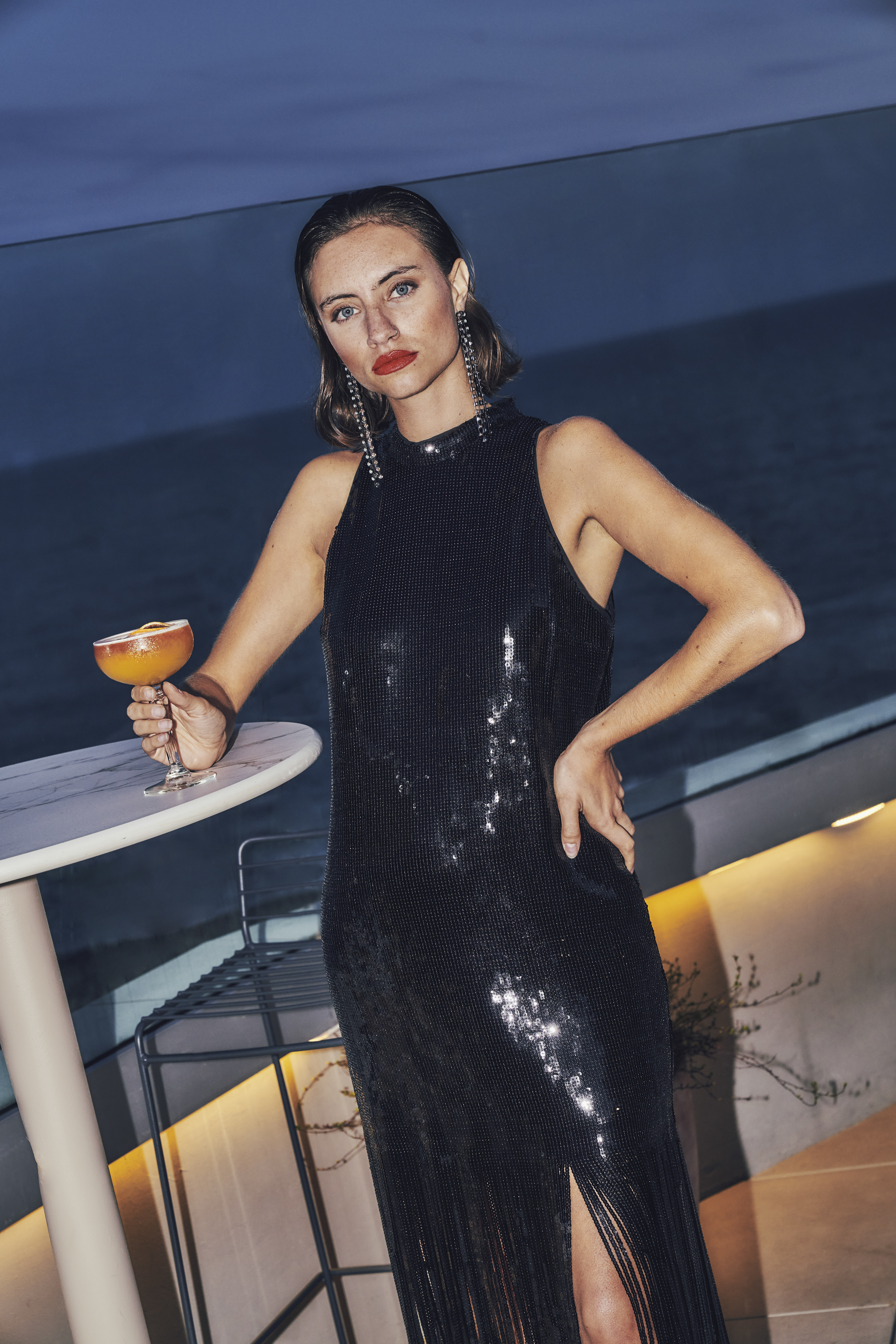 Model in black sequin high-neck dress holding a cocktail on a patio.