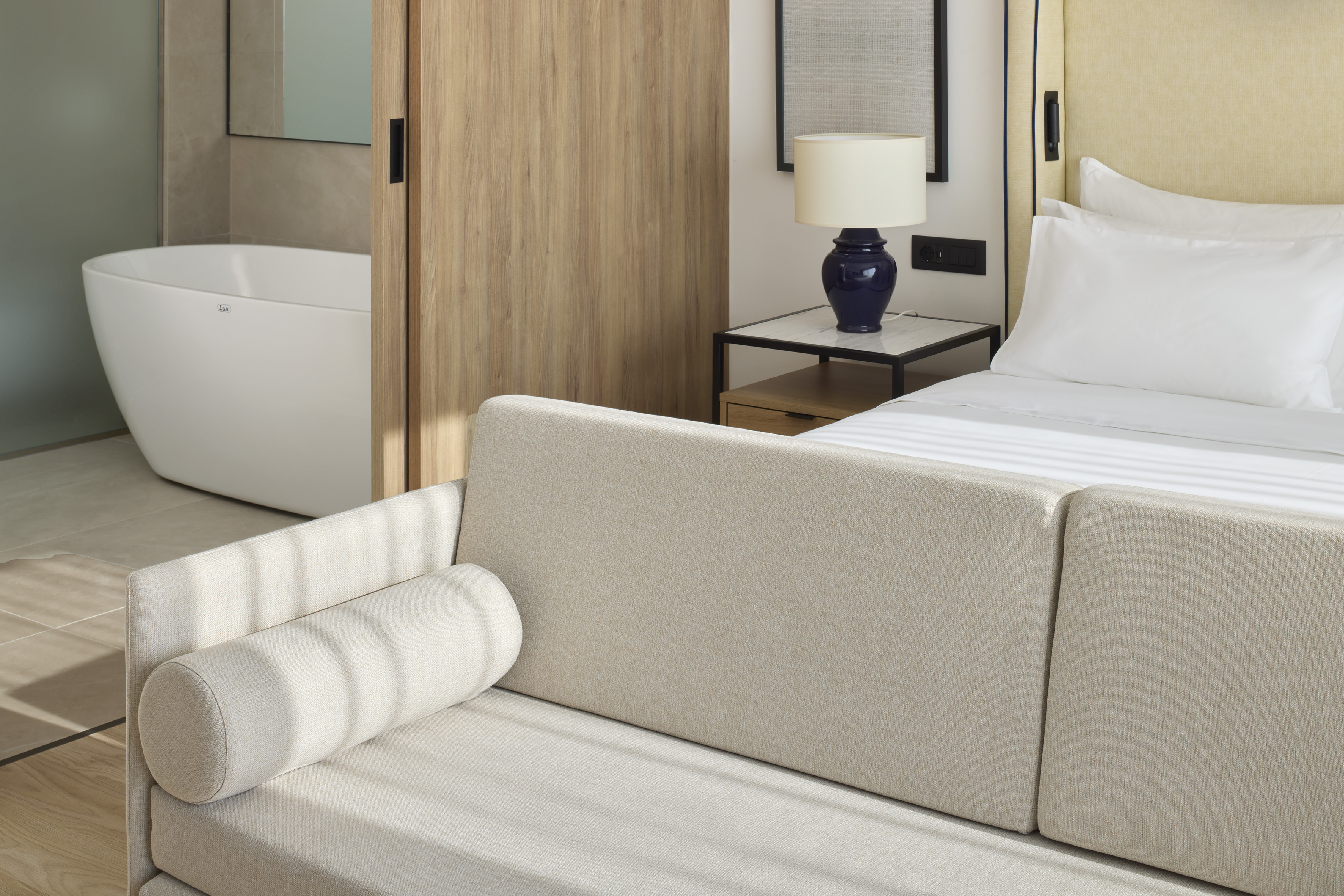 Modern hotel suite with bed, beige sofa, and luxury freestanding bathtub.