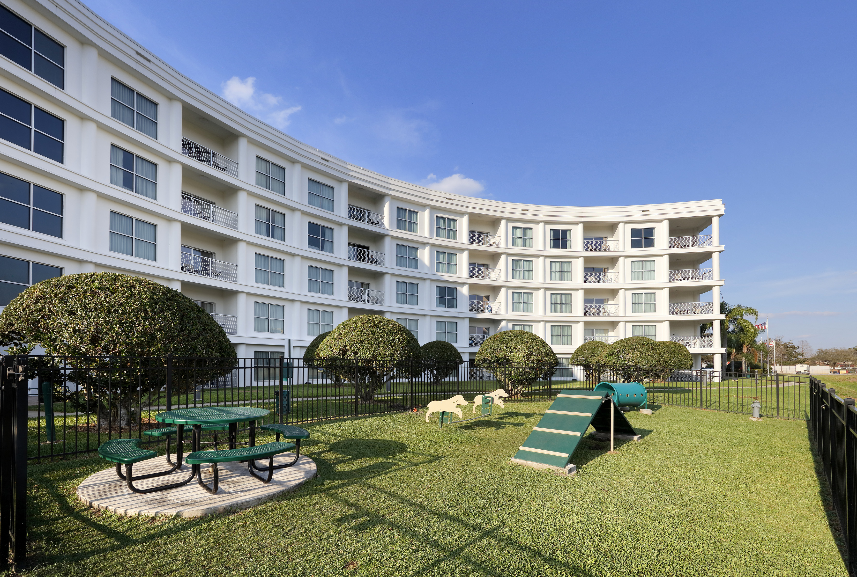 Curved white hotel building with pet park, picnic area, green bushes, and blue sky.