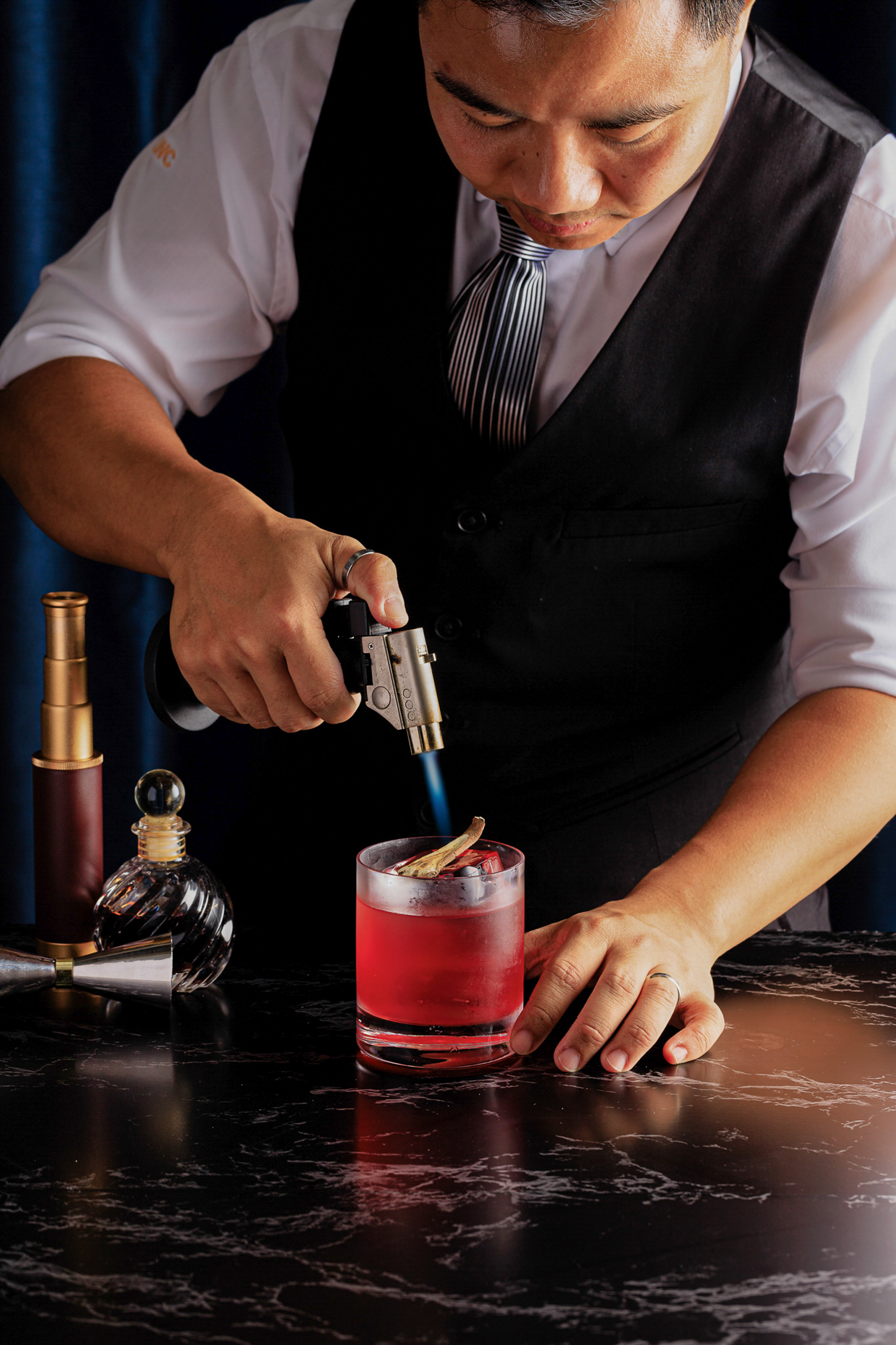 Bartender torching a red cocktail's garnish with a blue flame on a bar counter.