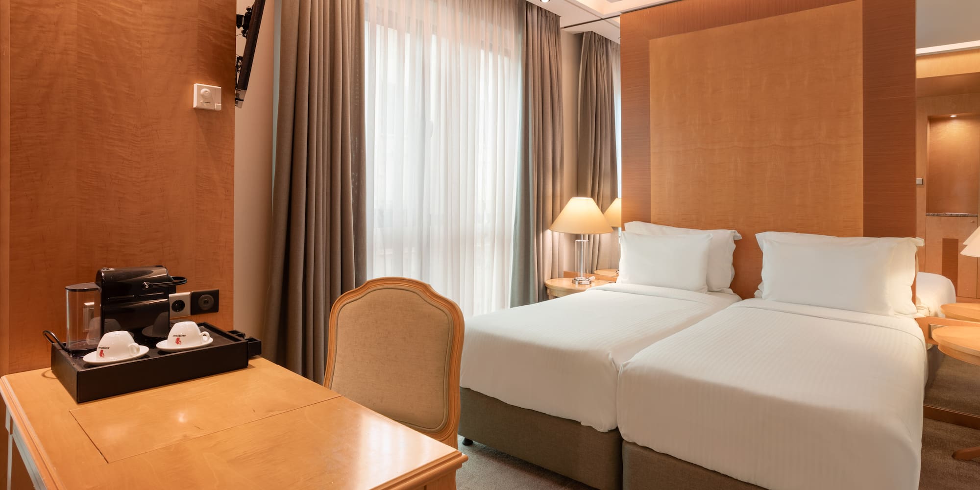 Hotel room with twin beds, wooden desk, coffee maker, TV, and large window.