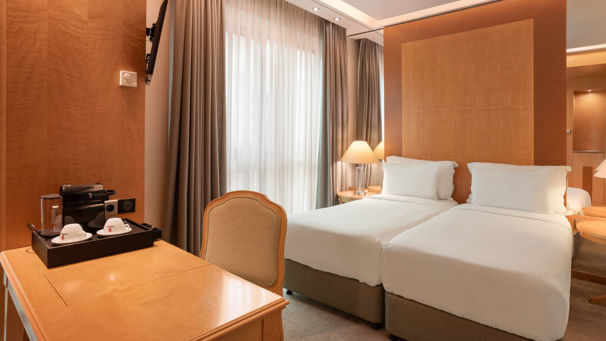 Hotel room with twin beds, wooden desk, coffee maker, TV, and large window.