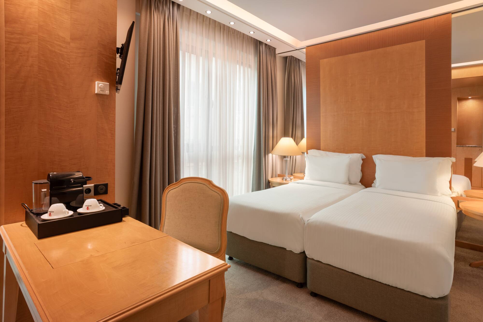 Hotel room with twin beds, wooden desk, coffee maker, TV, and large window.