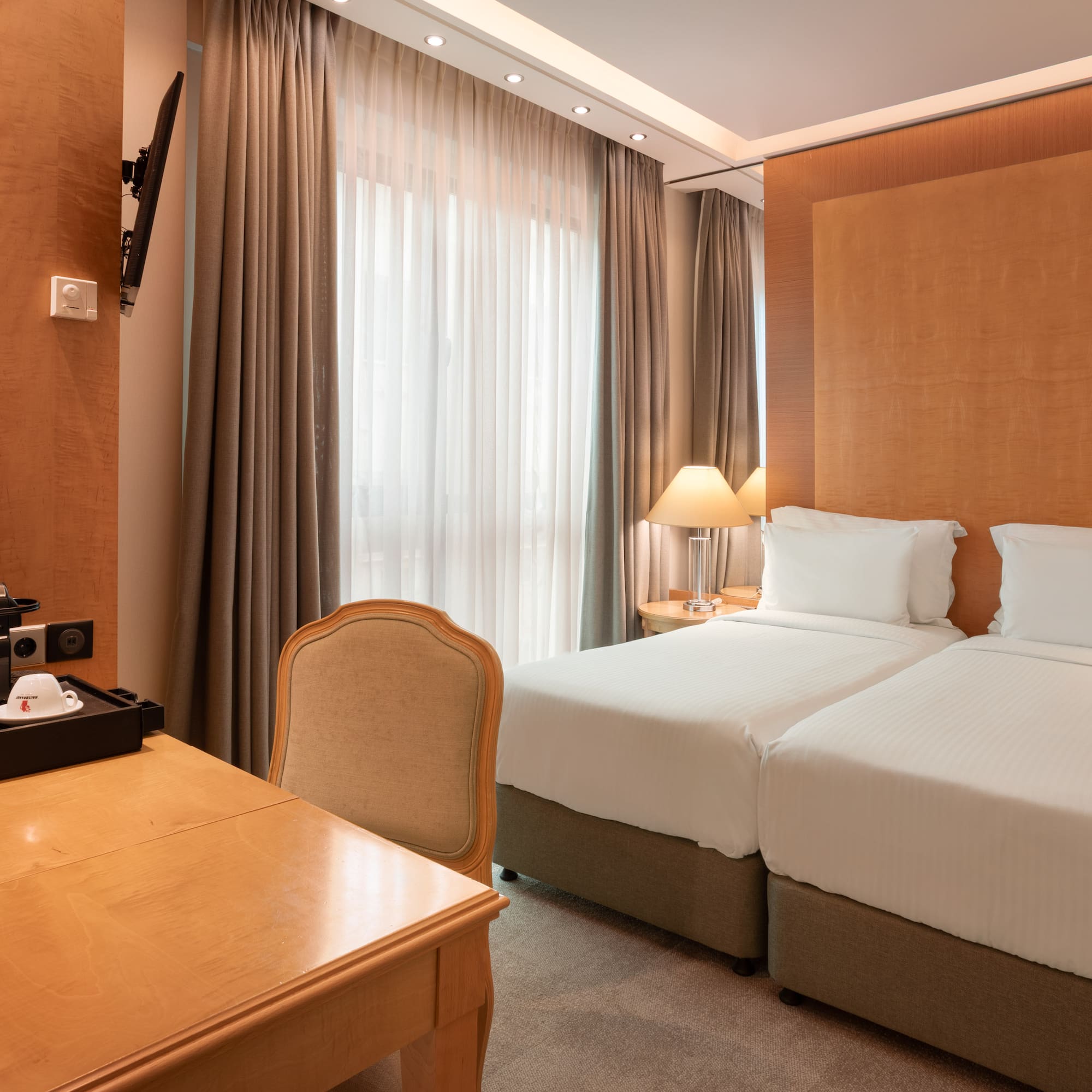 Hotel room with twin beds, wooden desk, coffee maker, TV, and large window.