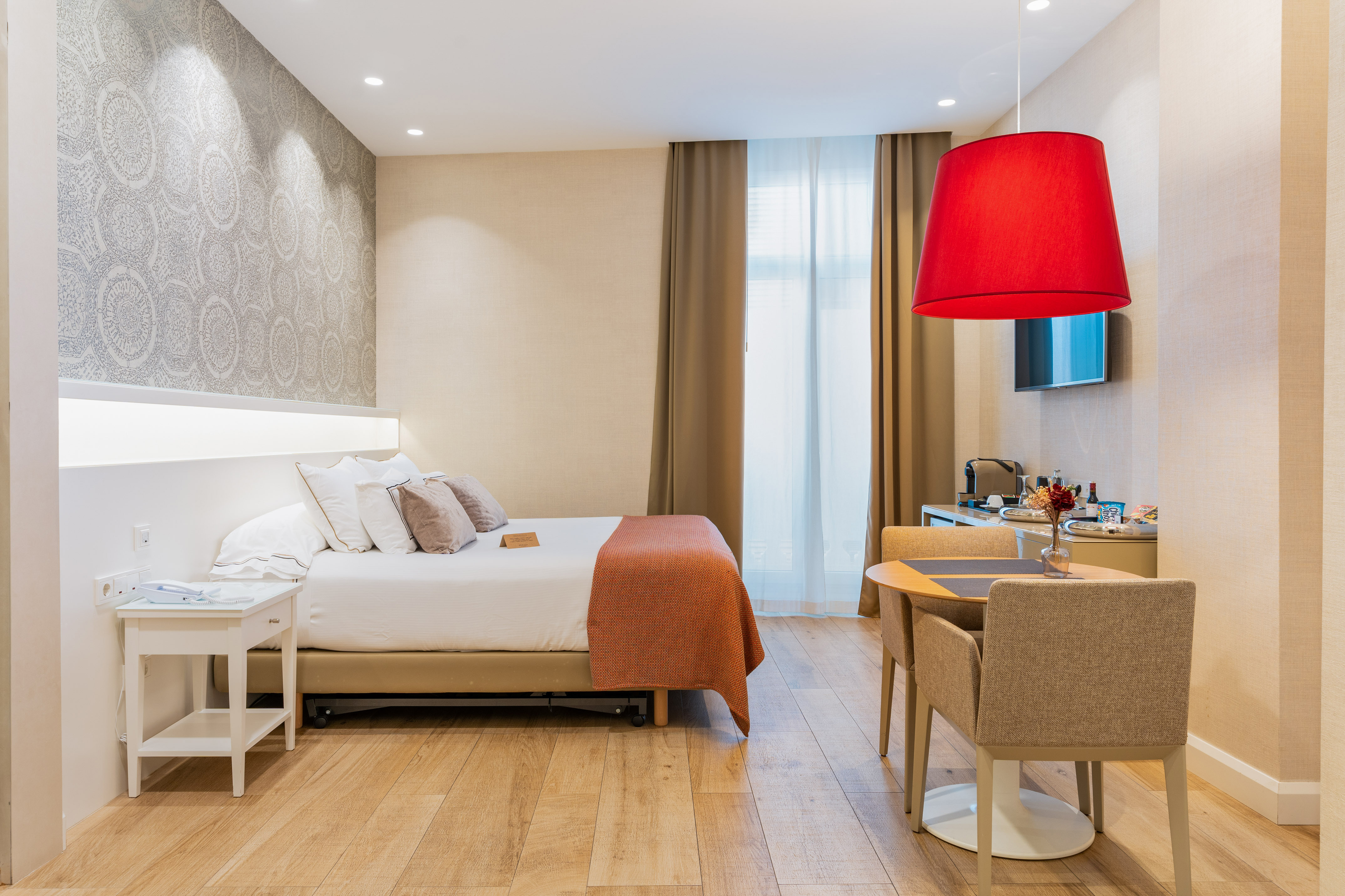 Modern hotel room with queen bed, dining area, wood floor, and bold red lamp.