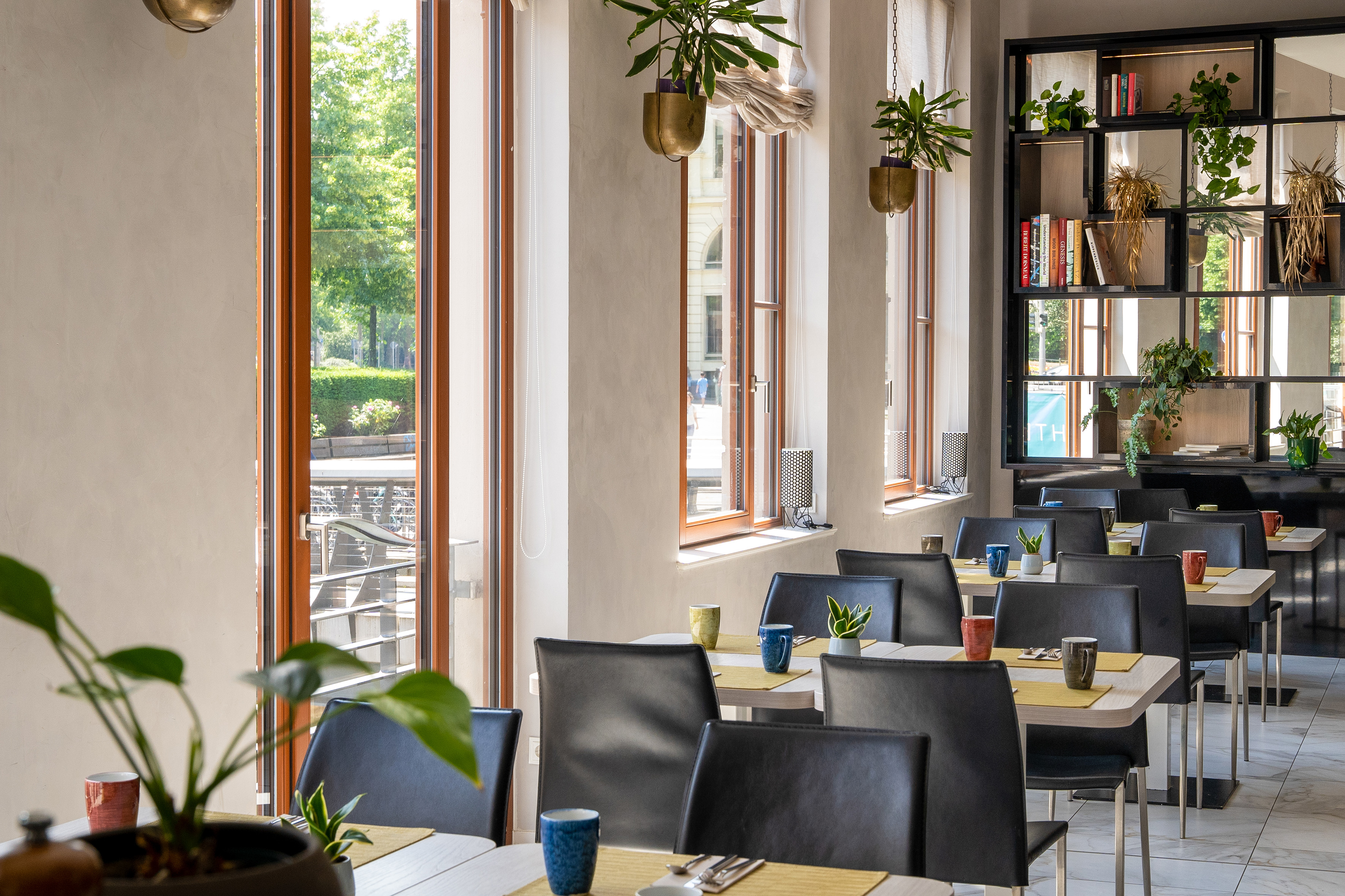 Modern restaurant dining room with tables, black chairs, large windows, and plants.