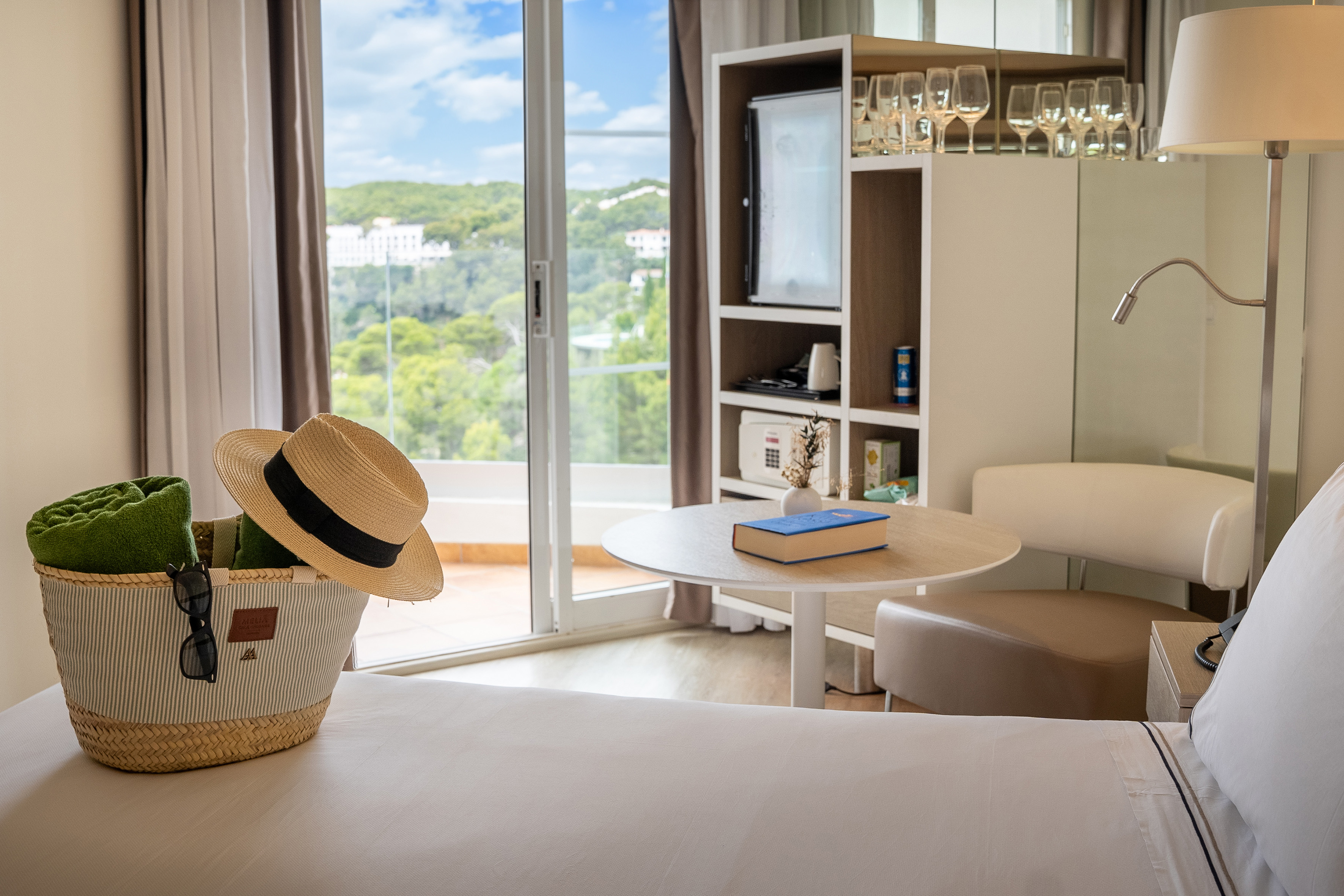 Modern hotel room with beach bag, straw hat, and sunny balcony overlooking green hills.