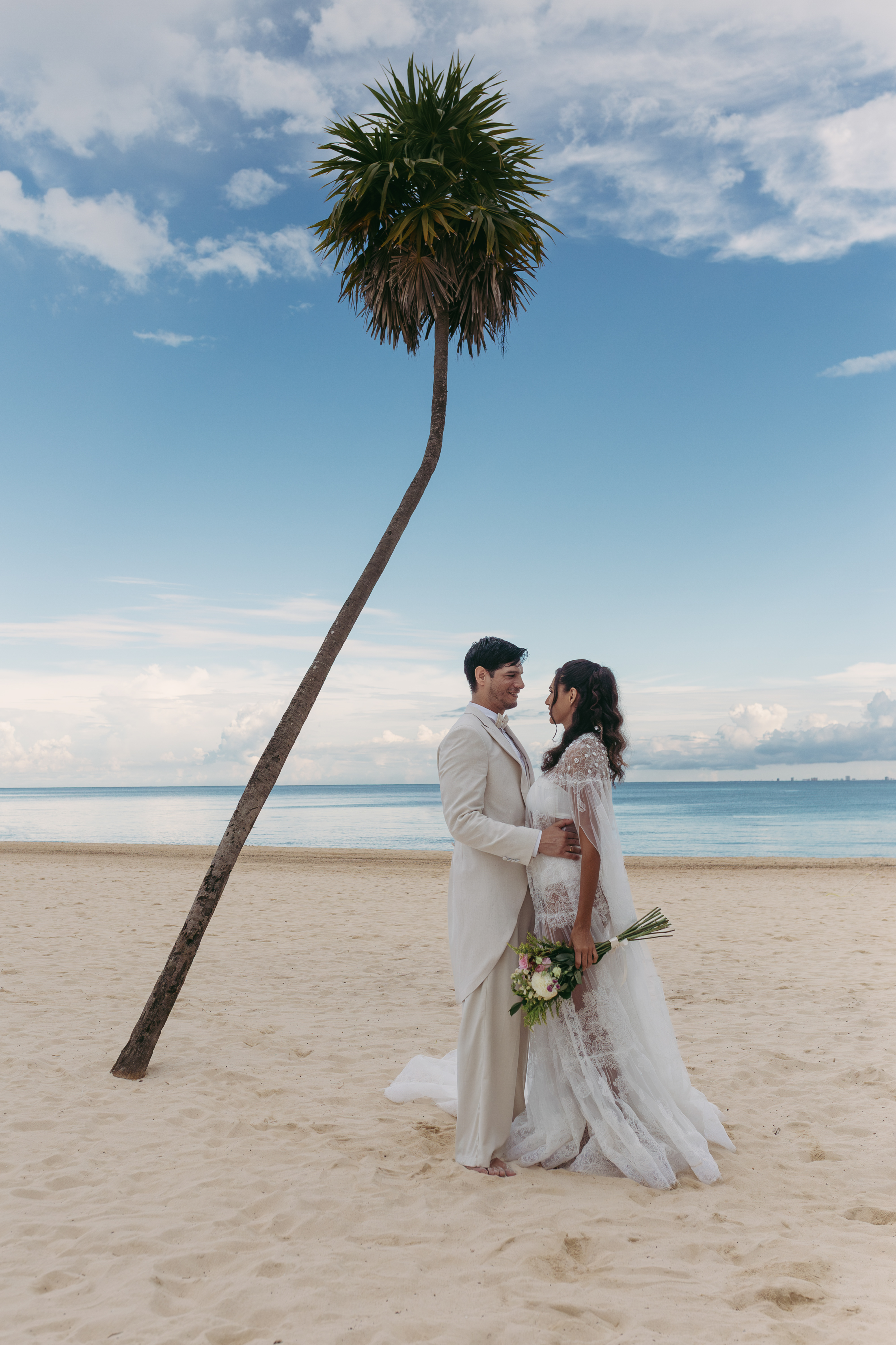 Bride and groom embrace on a tropical beach under a leaning palm tree.