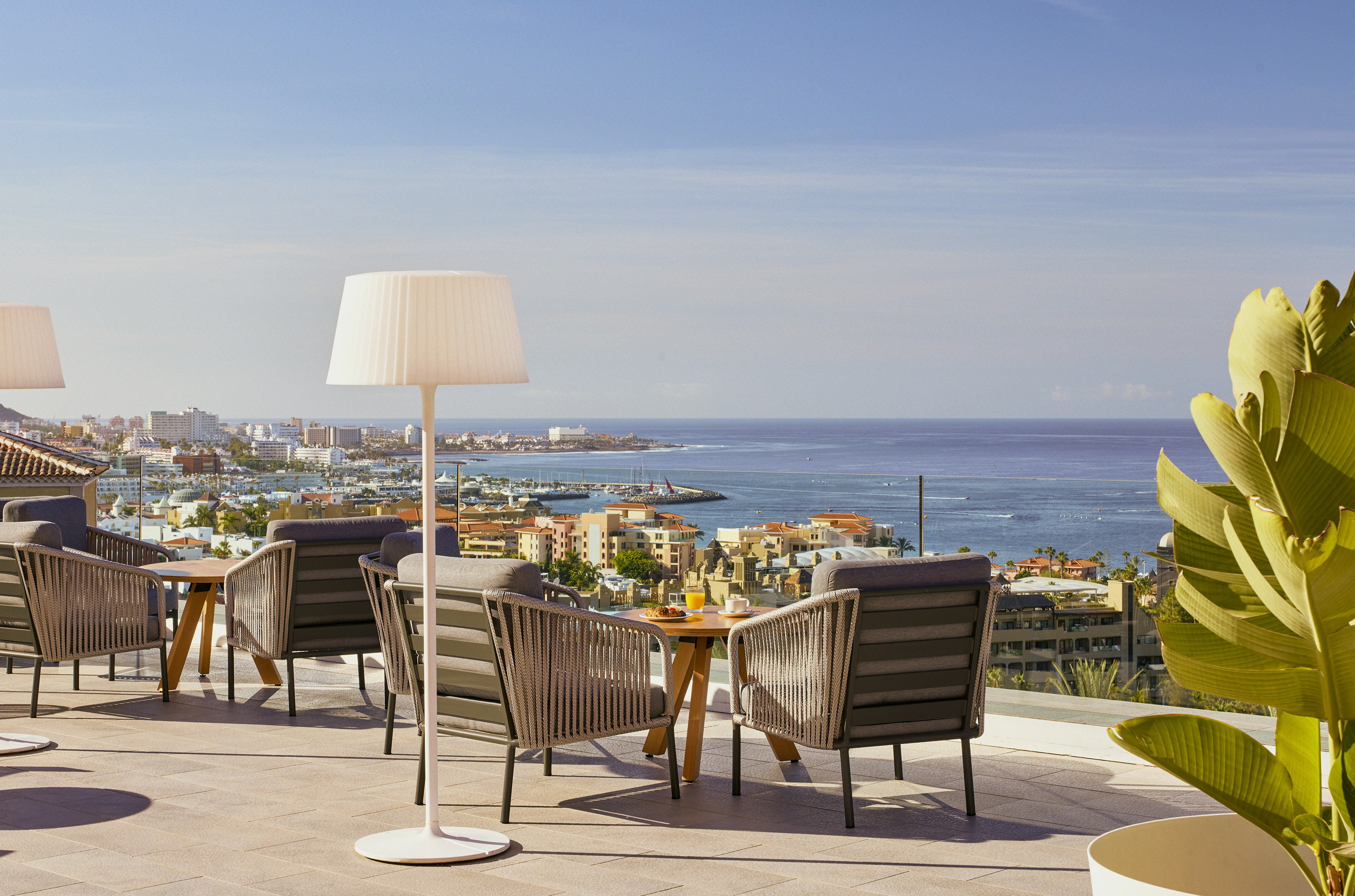 Sunny terrace overlooking a vibrant coastal city, ocean, with outdoor dining.