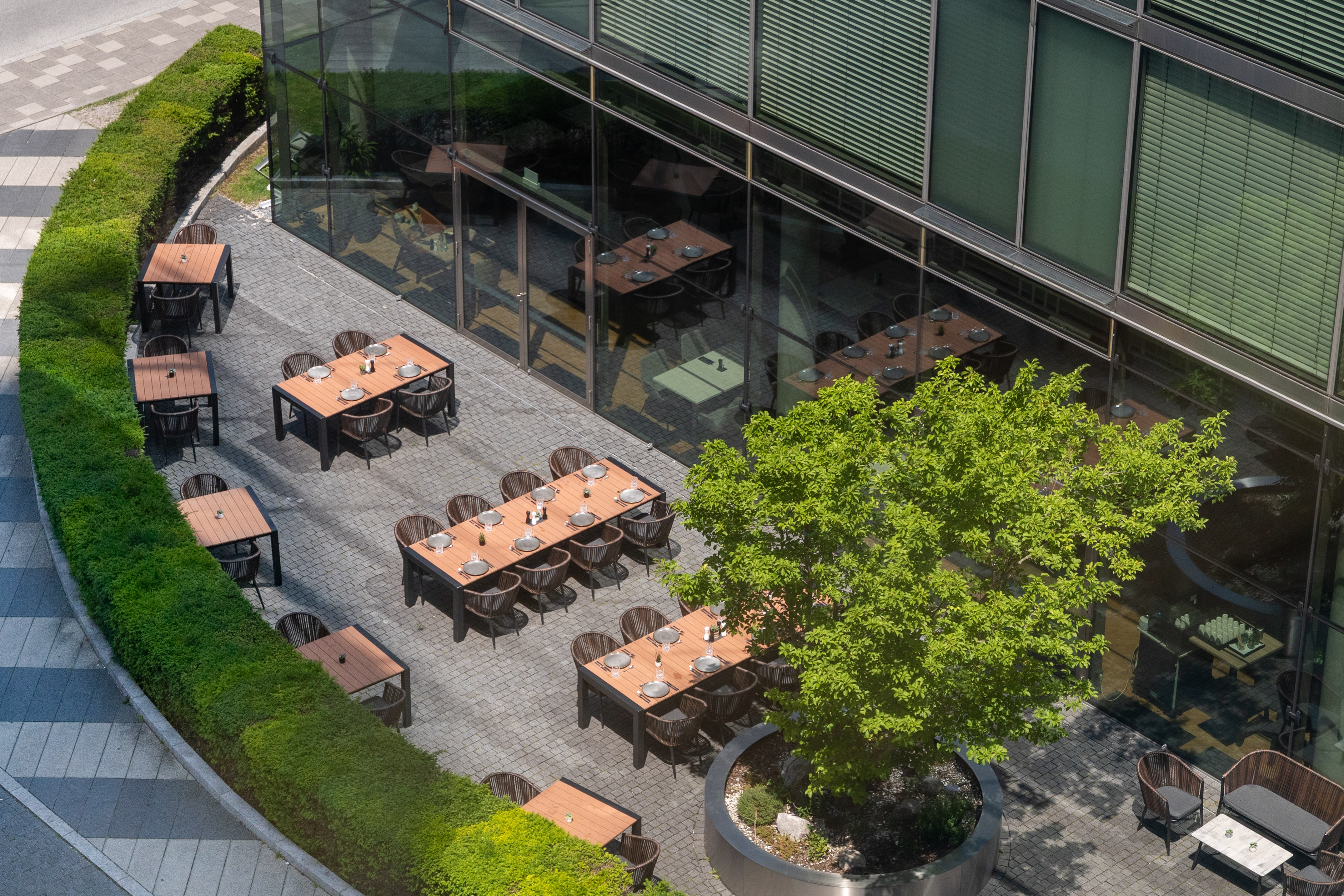 Modern outdoor restaurant patio with dining tables, green tree, and glass building.