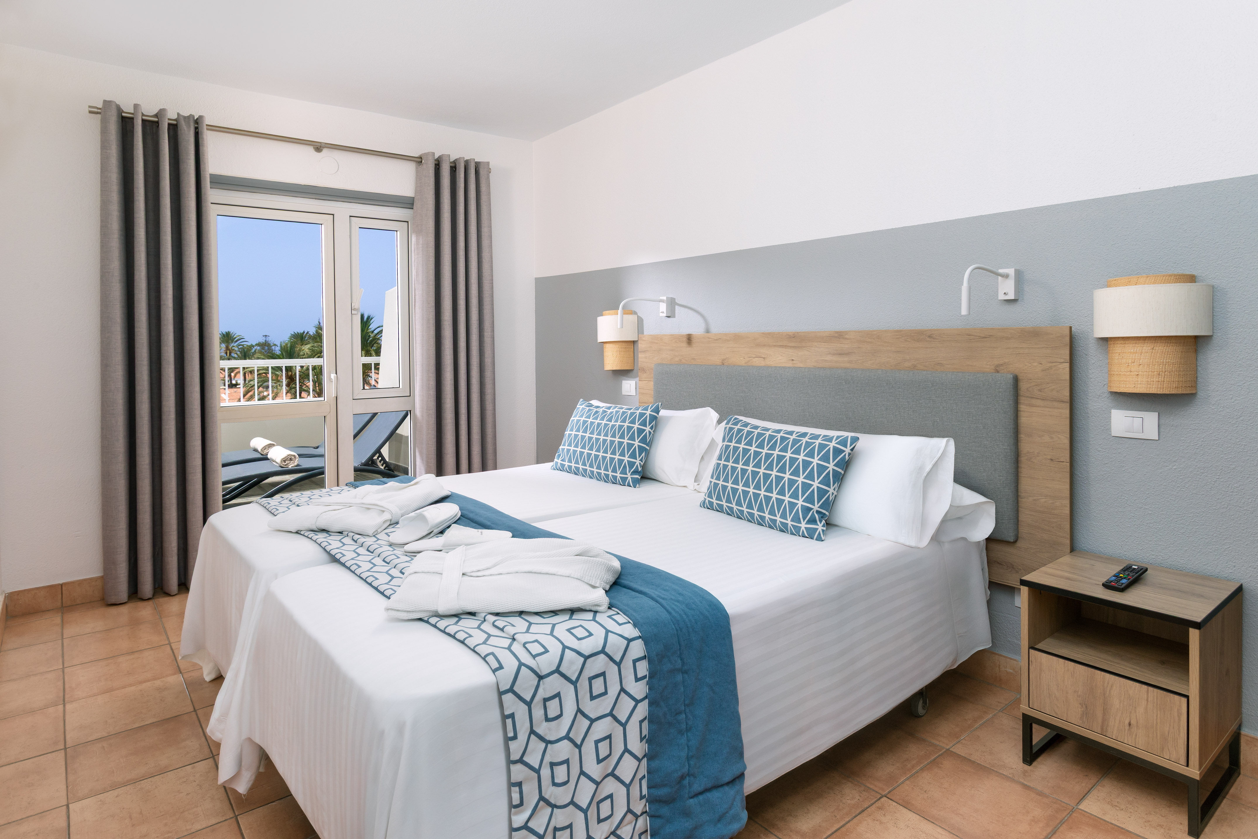 Modern hotel room with twin beds, blue decor, balcony, and tropical view.