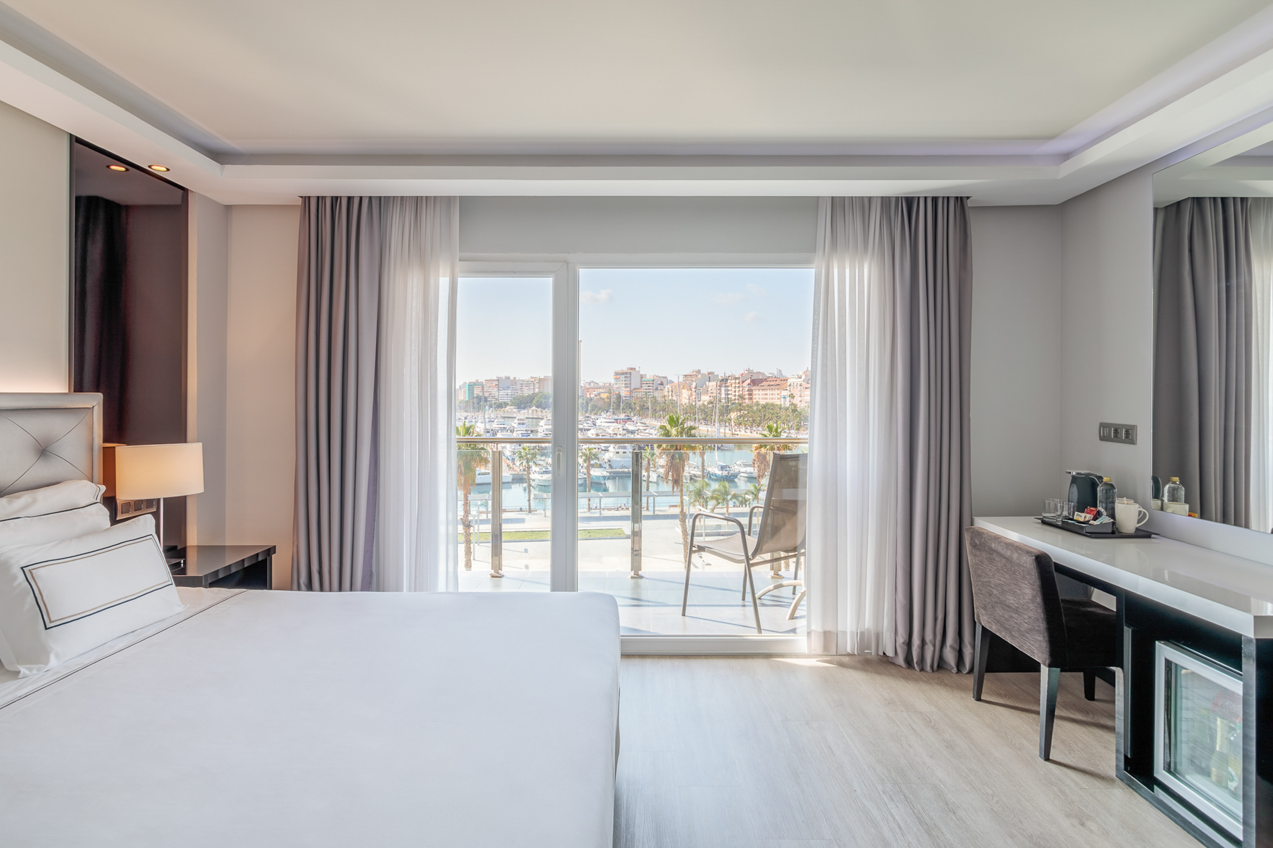 Modern hotel room with marina view balcony, white bed, and desk.