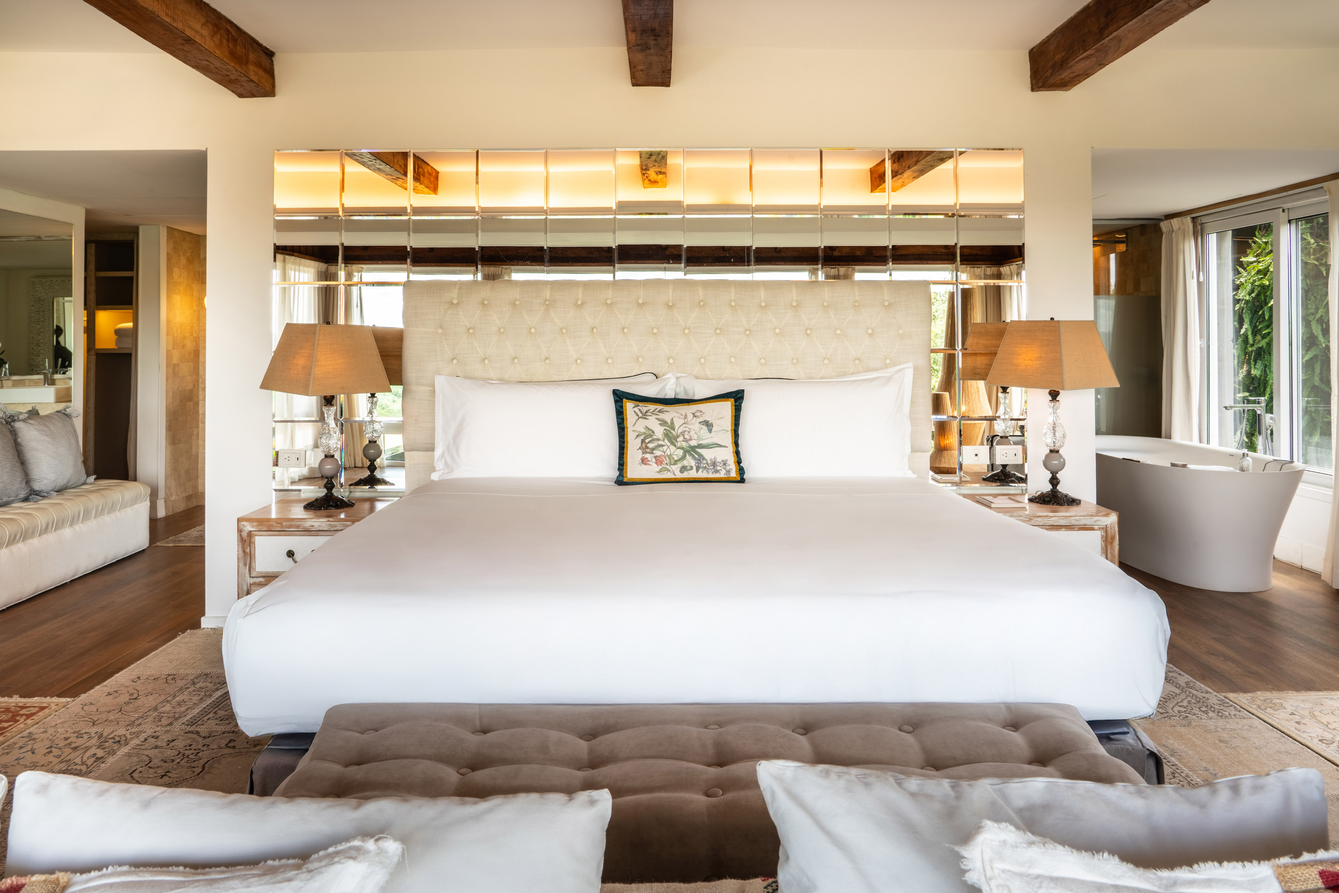 King bedroom with tufted headboard, mirrored wall, wood beams, and freestanding tub.