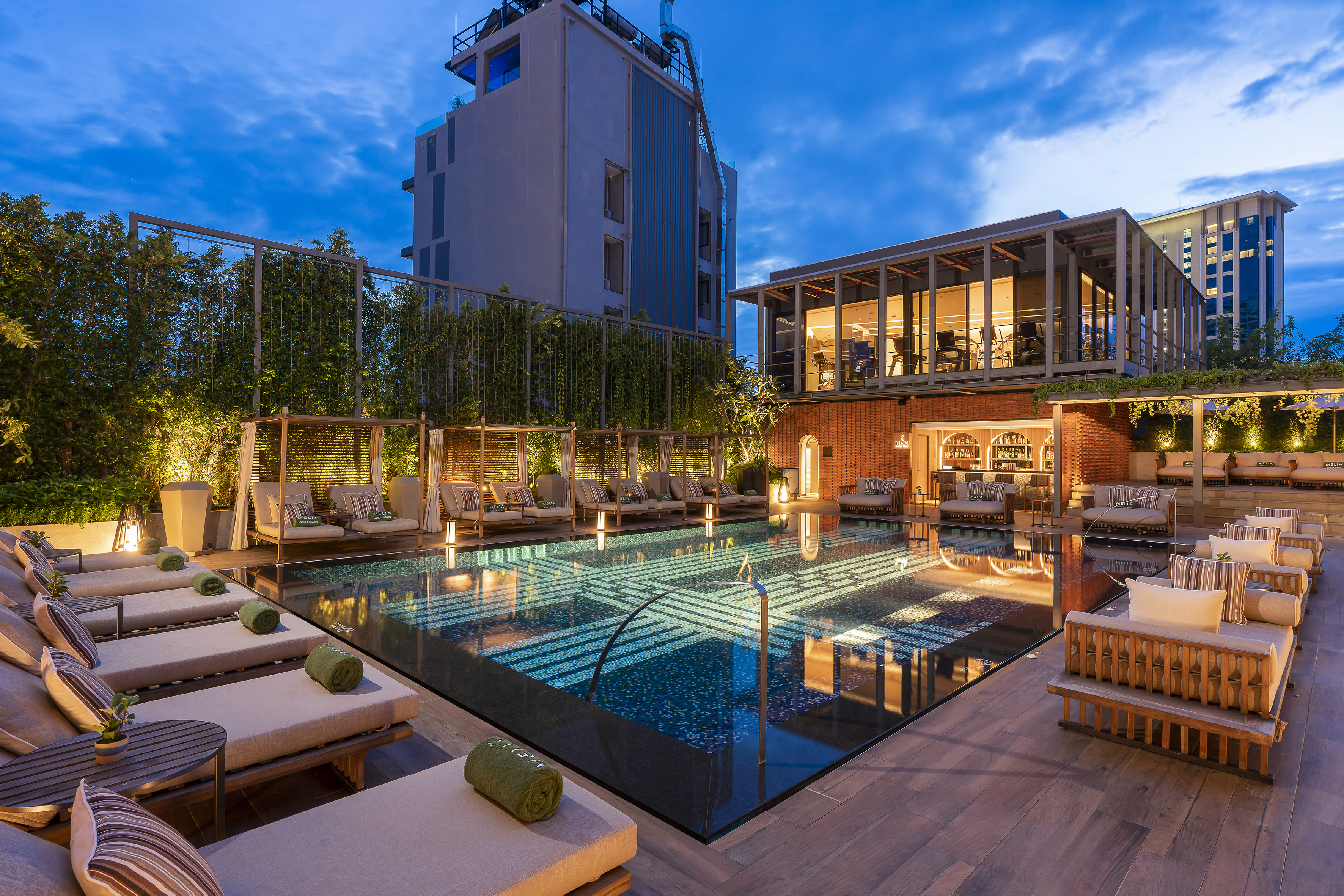 Evening rooftop hotel pool with loungers, bar, and city skyline.