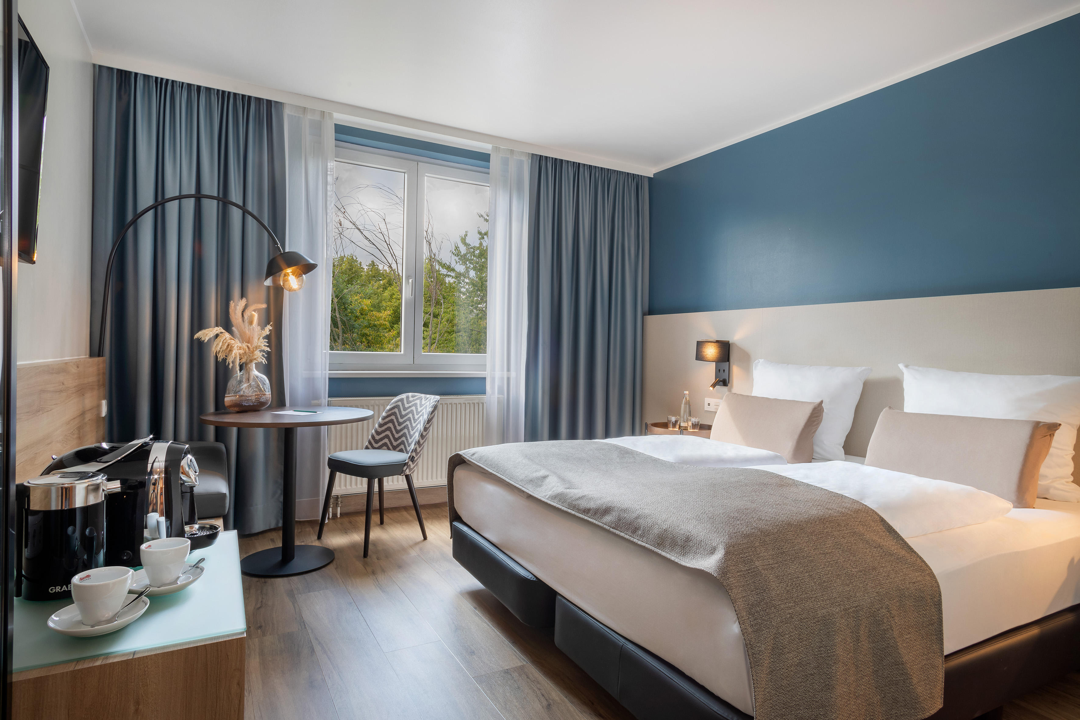 Stylish hotel room: double bed, blue wall, forest view, coffee machine.