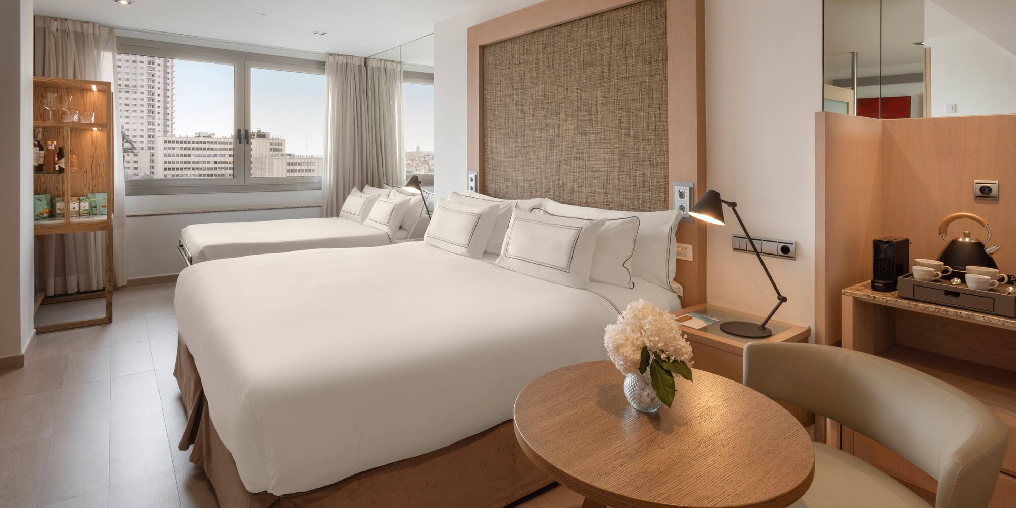 Modern hotel room with double bed, single bed, city view, and coffee station.