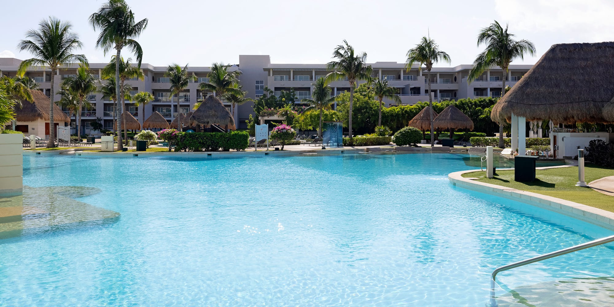 Large blue hotel swimming pool surrounded by palm trees and tropical huts.