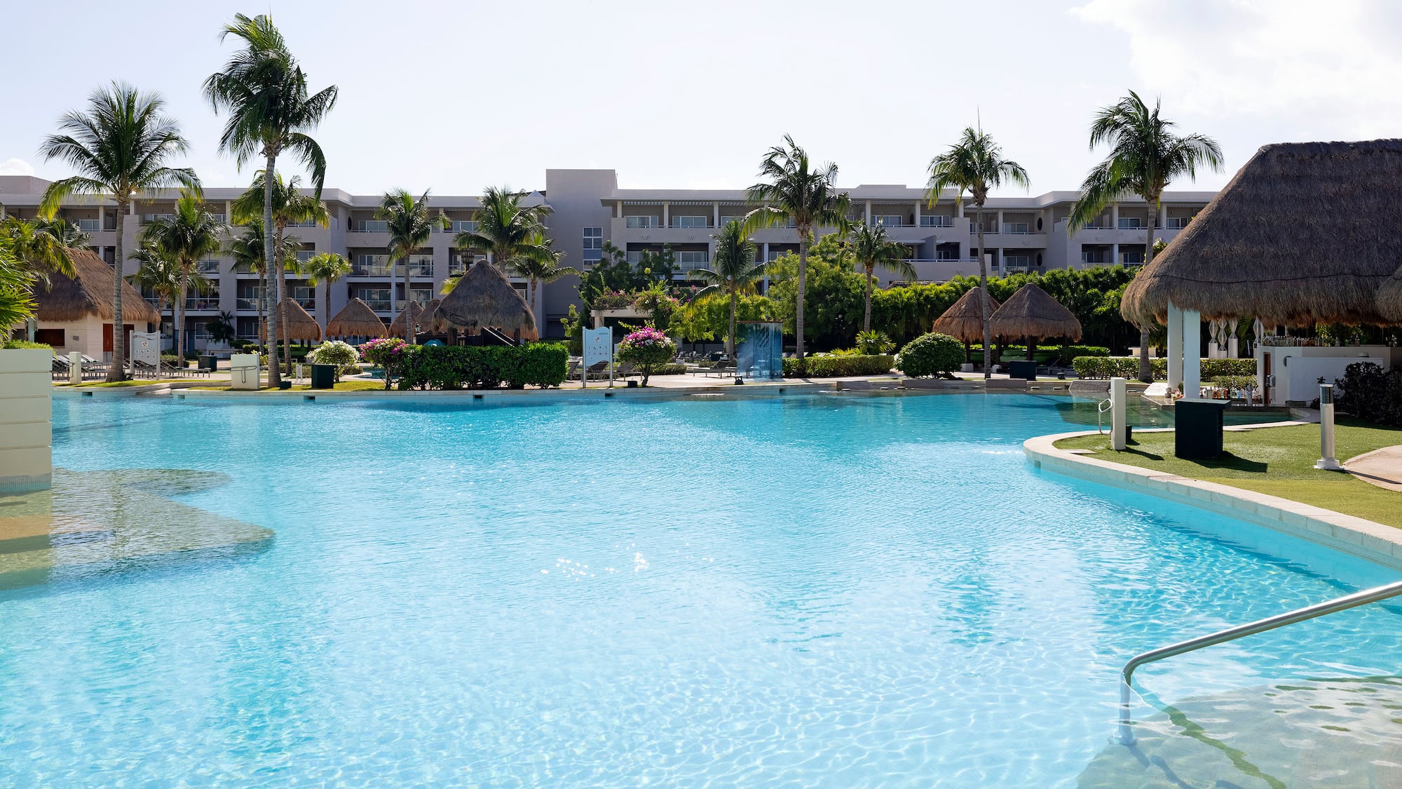 Large blue hotel swimming pool surrounded by palm trees and tropical huts.