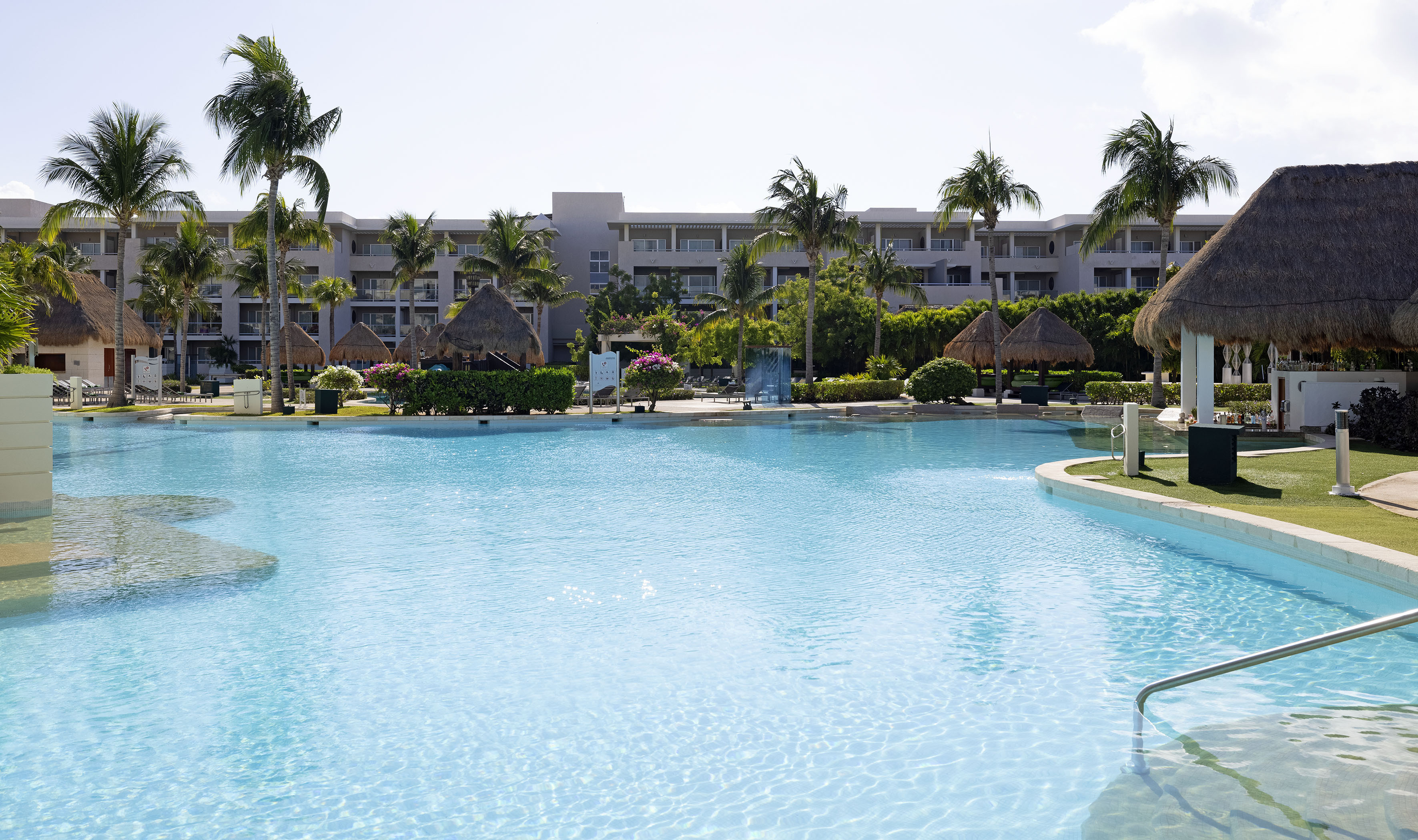 Large blue hotel swimming pool surrounded by palm trees and tropical huts.