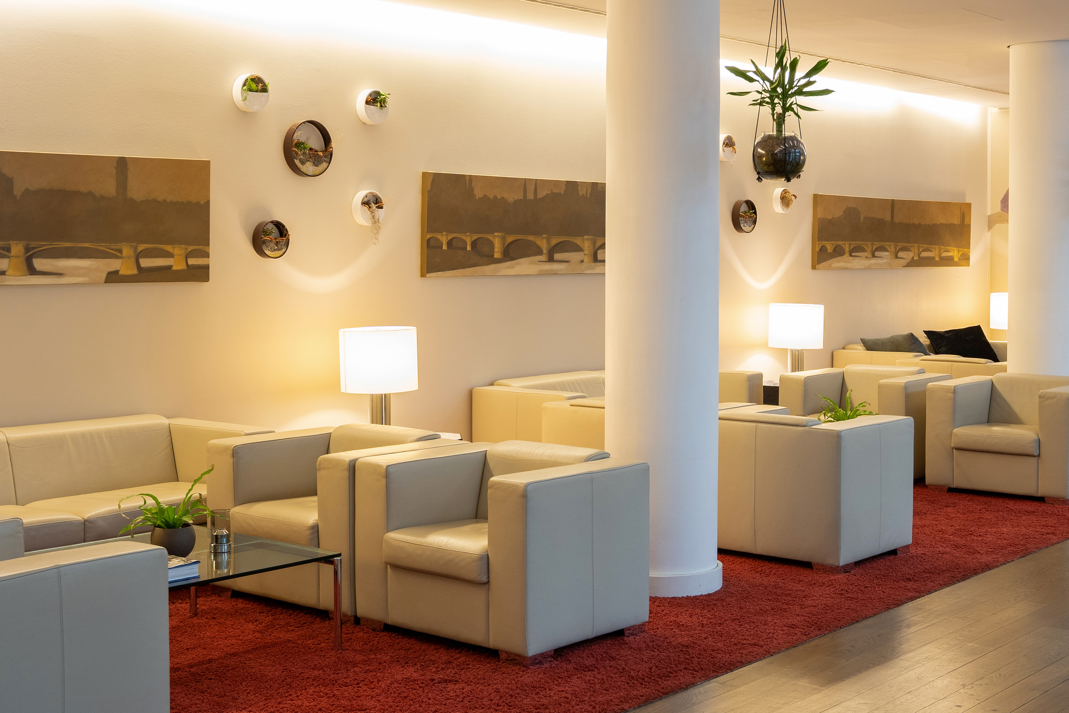 Modern hotel lounge with cream sofas, red carpet, bridge artwork, and plants.